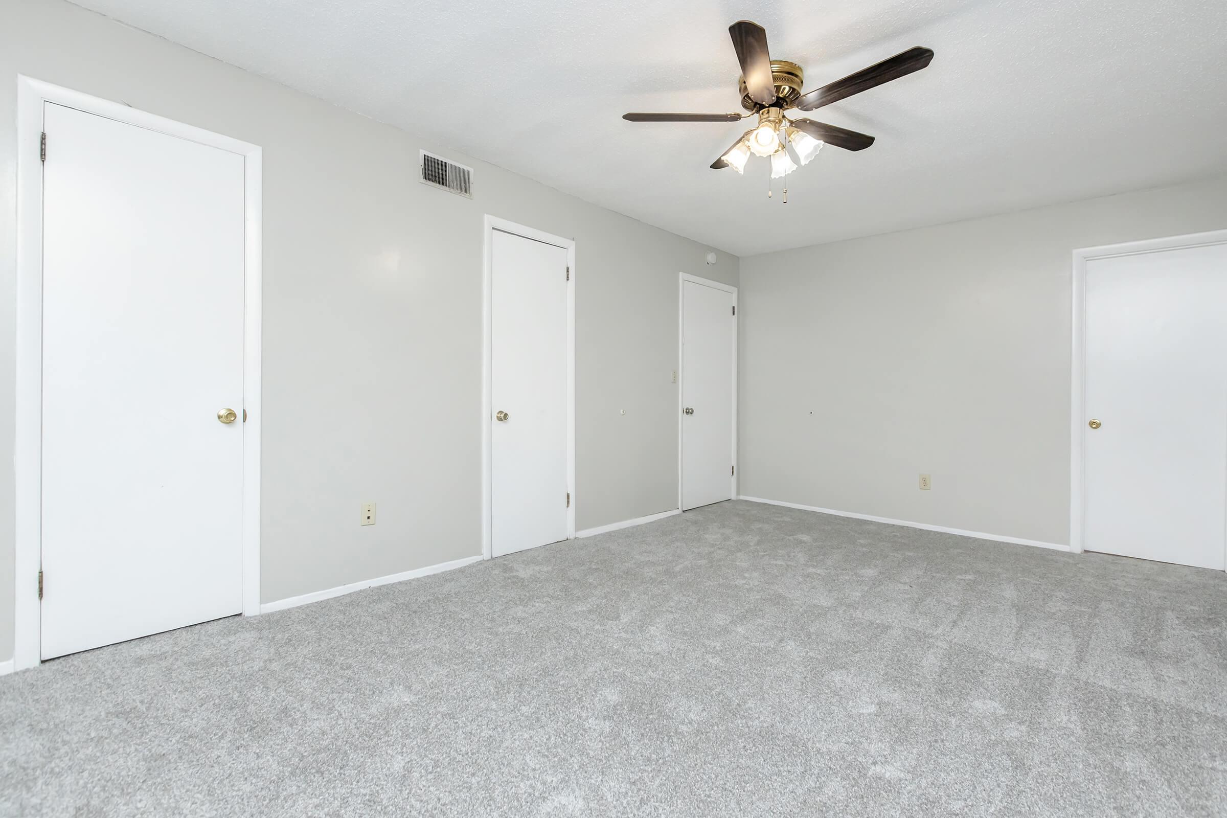 A spacious, empty room with light gray walls and plush gray carpeting. The room features a ceiling fan with five blades, and three white doors on the left and right walls, leading to other spaces. The overall atmosphere is bright and inviting, allowing for various decorating possibilities.