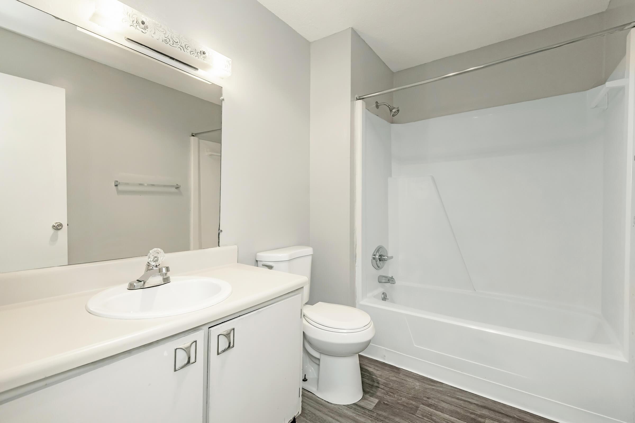 A modern bathroom featuring a white bathtub with a shower, a white sink with a mirror above, a toilet, and light gray walls. The flooring is a dark wood-like laminate, enhancing the contemporary design of the space.