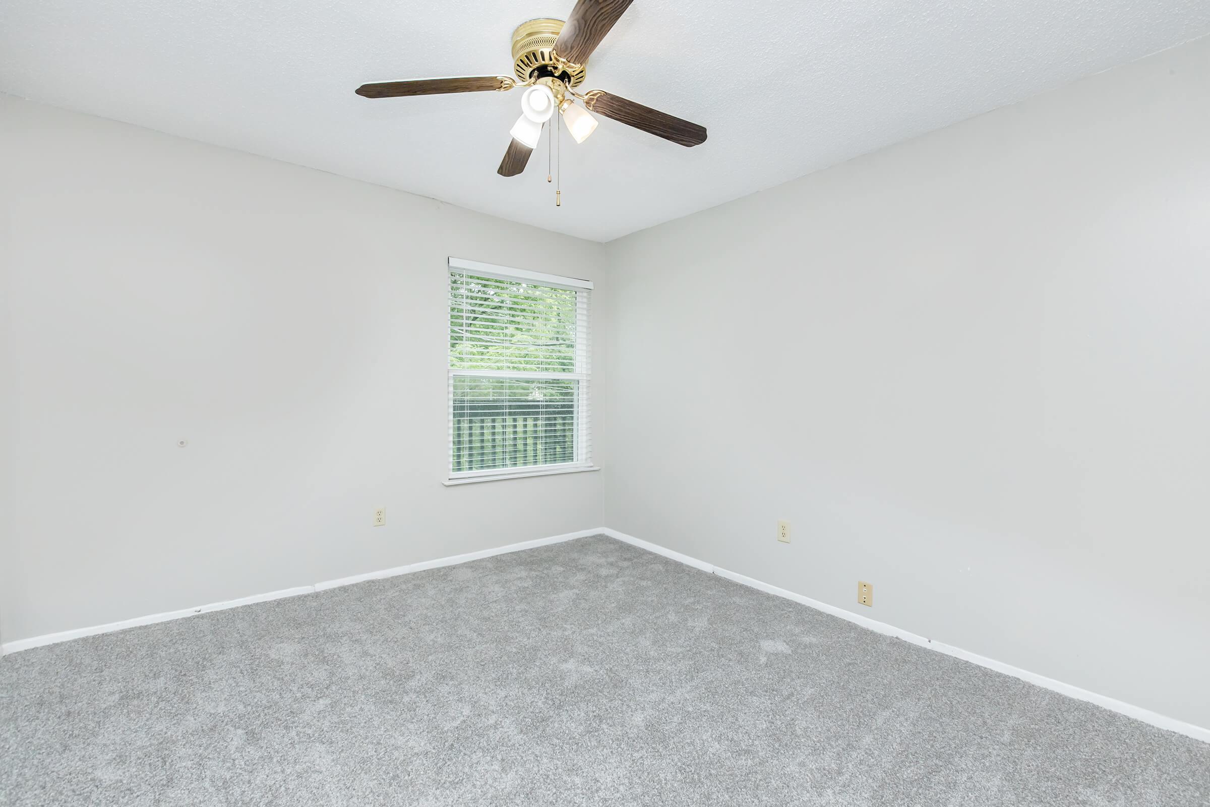 A vacant room featuring light gray carpet, a ceiling fan with three light fixtures, and a single window letting in natural light. The walls are painted a neutral color, and there are no furnishings, giving the space a spacious and airy feel.