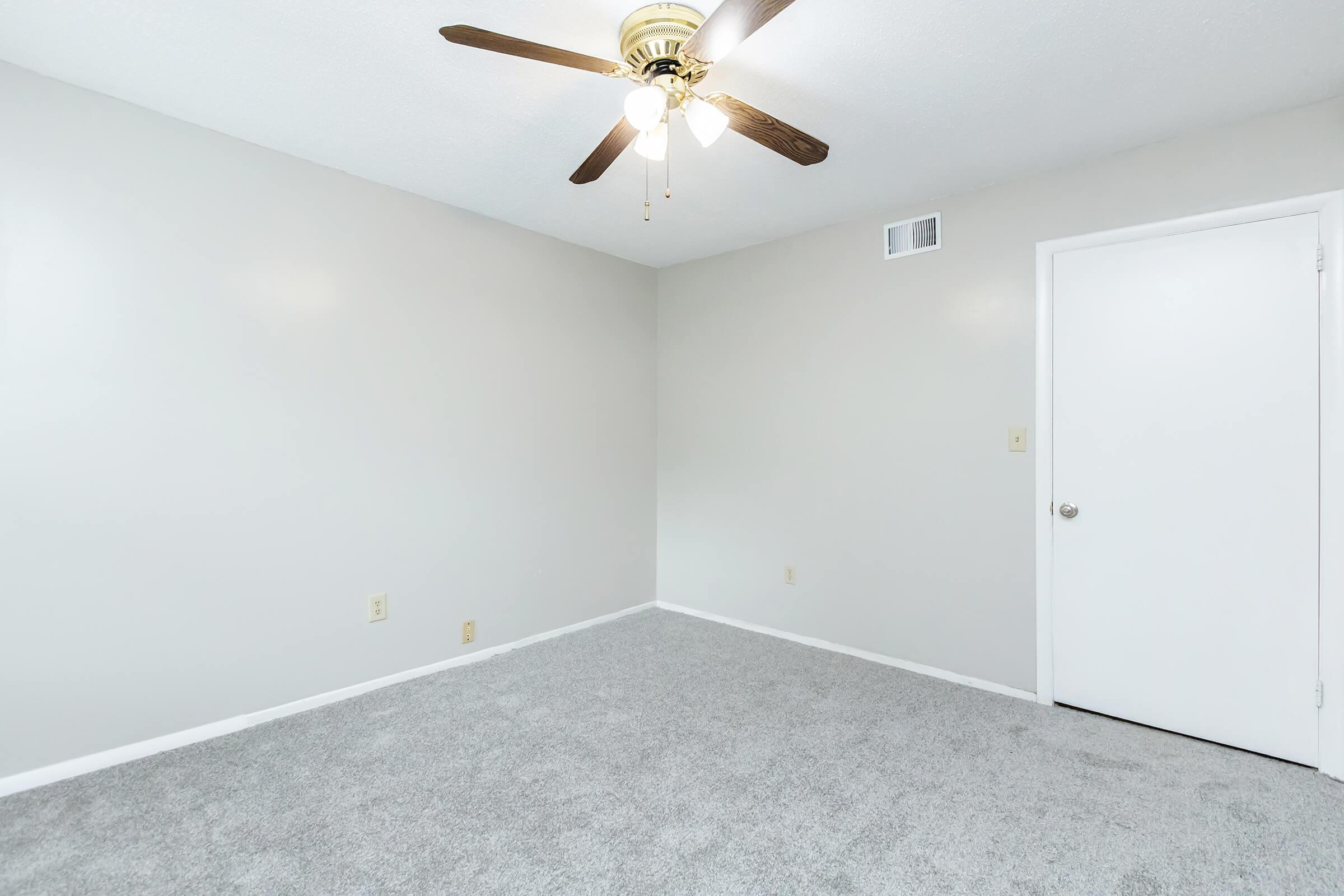 A spacious, empty room with light gray carpet and neutral-colored walls. A ceiling fan with wooden blades is visible, and there's a closed white door on the right side. The room has no furniture, creating a bright and airy atmosphere.