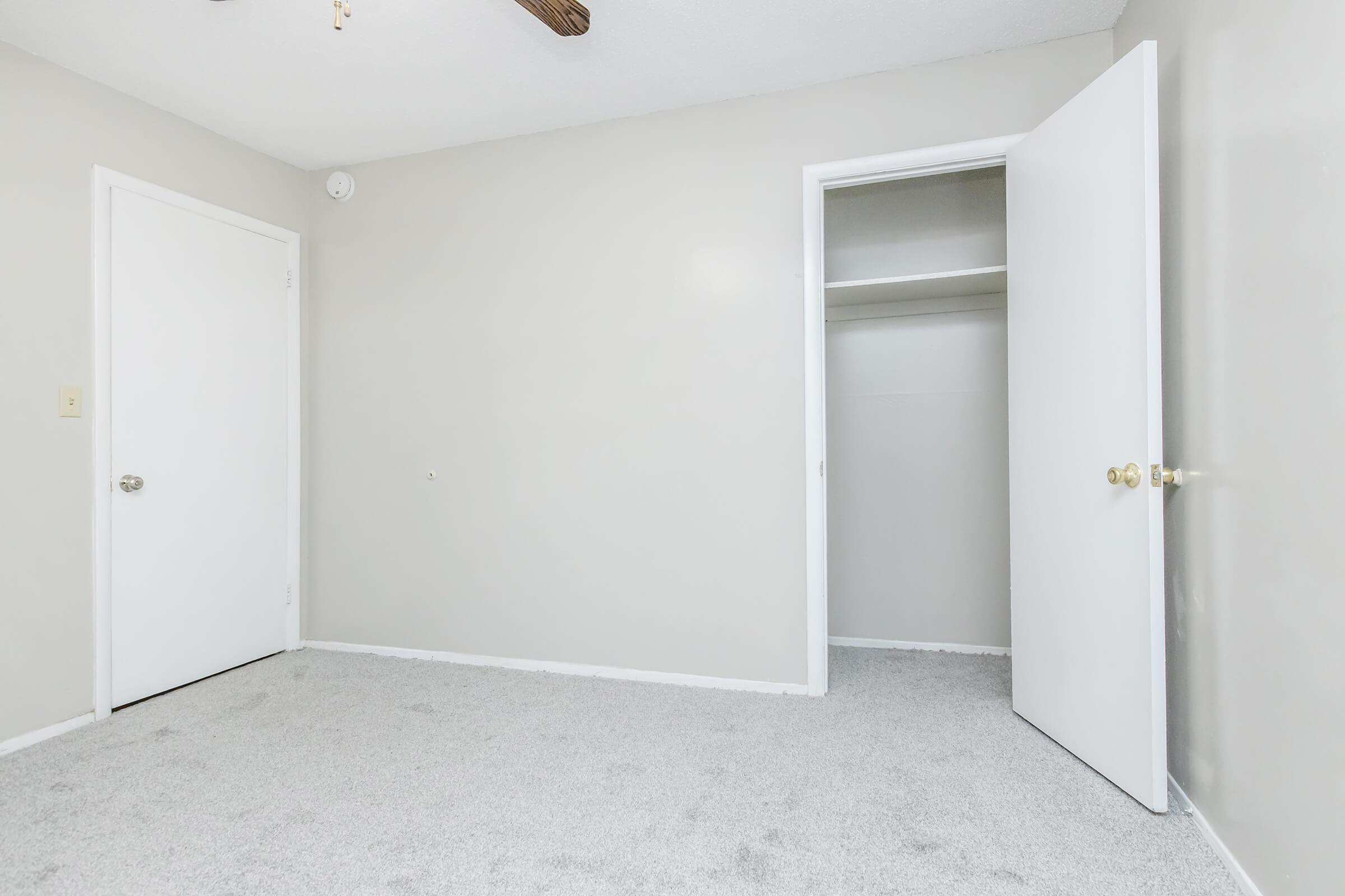 Empty room with light gray walls and carpet. A white door on the left leads to another space, while an open closet is visible on the right. A ceiling fan hangs from the ceiling, adding to the simplicity of the room's design. Natural light illuminates the space, creating a bright atmosphere.