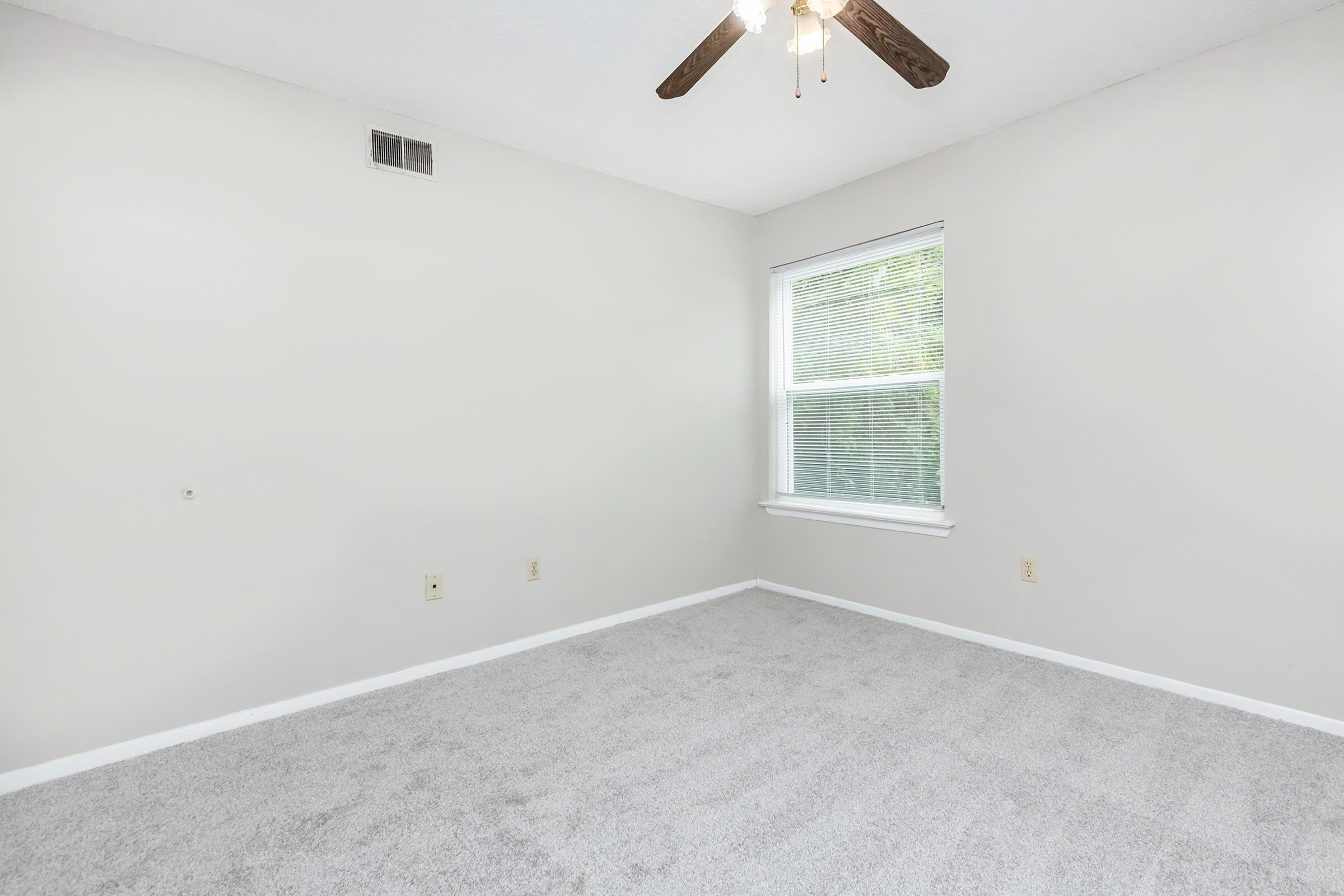 Empty room with light gray carpet and pale beige walls. A small window with blinds allows natural light, and a ceiling fan with wooden blades is visible. No furniture or decor is present, creating a spacious and minimalistic atmosphere.