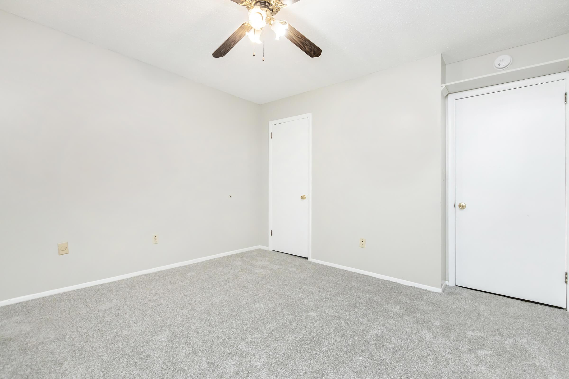 Empty room with light beige walls and a textured grey carpet. A ceiling fan with light fixtures is visible in the center. There are two white doors, one closet and one leading to another room, both closed. The space is well-lit and has a clean, spacious feel.