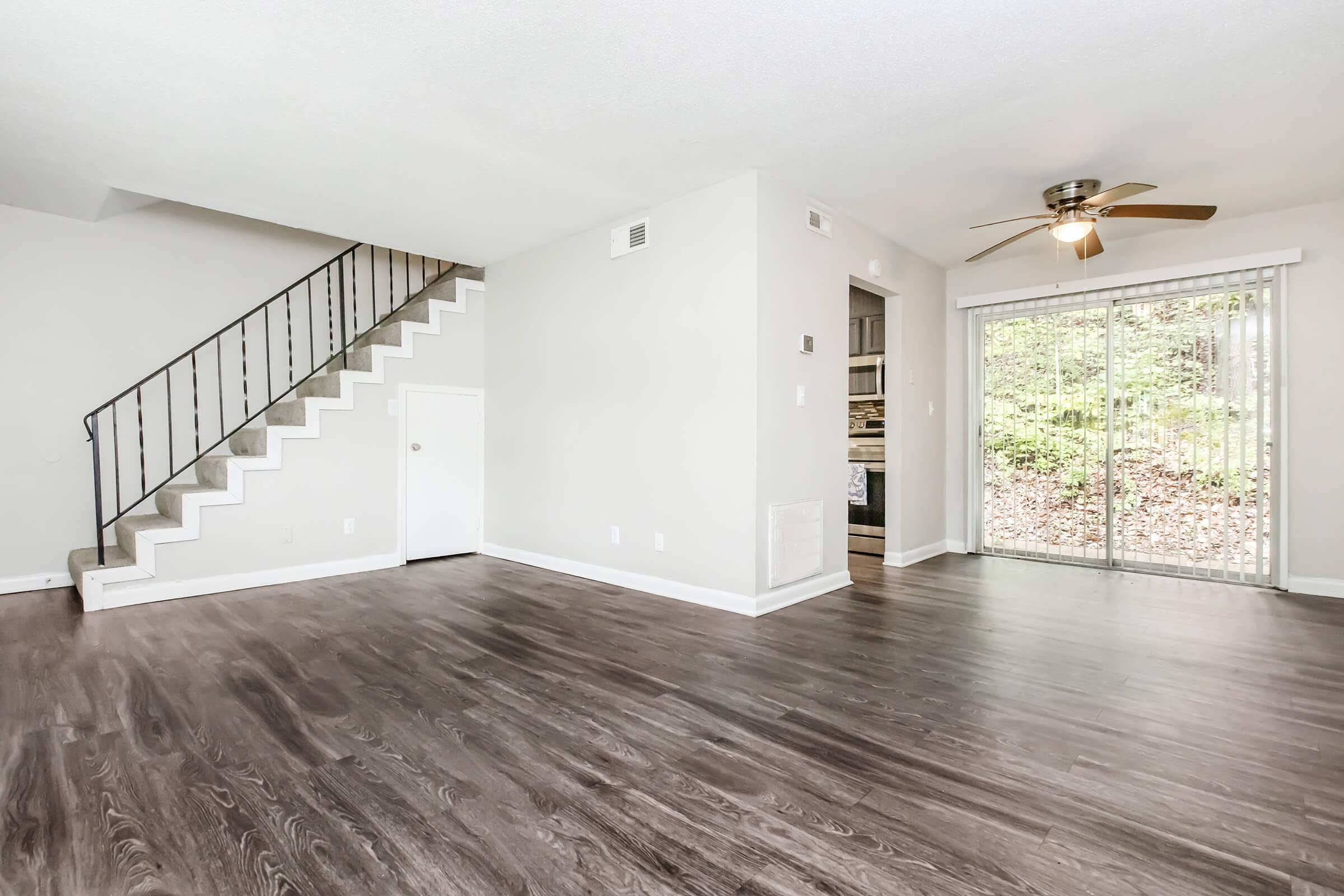 A spacious living room area featuring a light-colored wall, wood-like flooring, and a ceiling fan. A staircase leads to the upper level on the left, while a sliding glass door on the right opens to a view of greenery outside. The space is bright and inviting, with a minimalistic design.