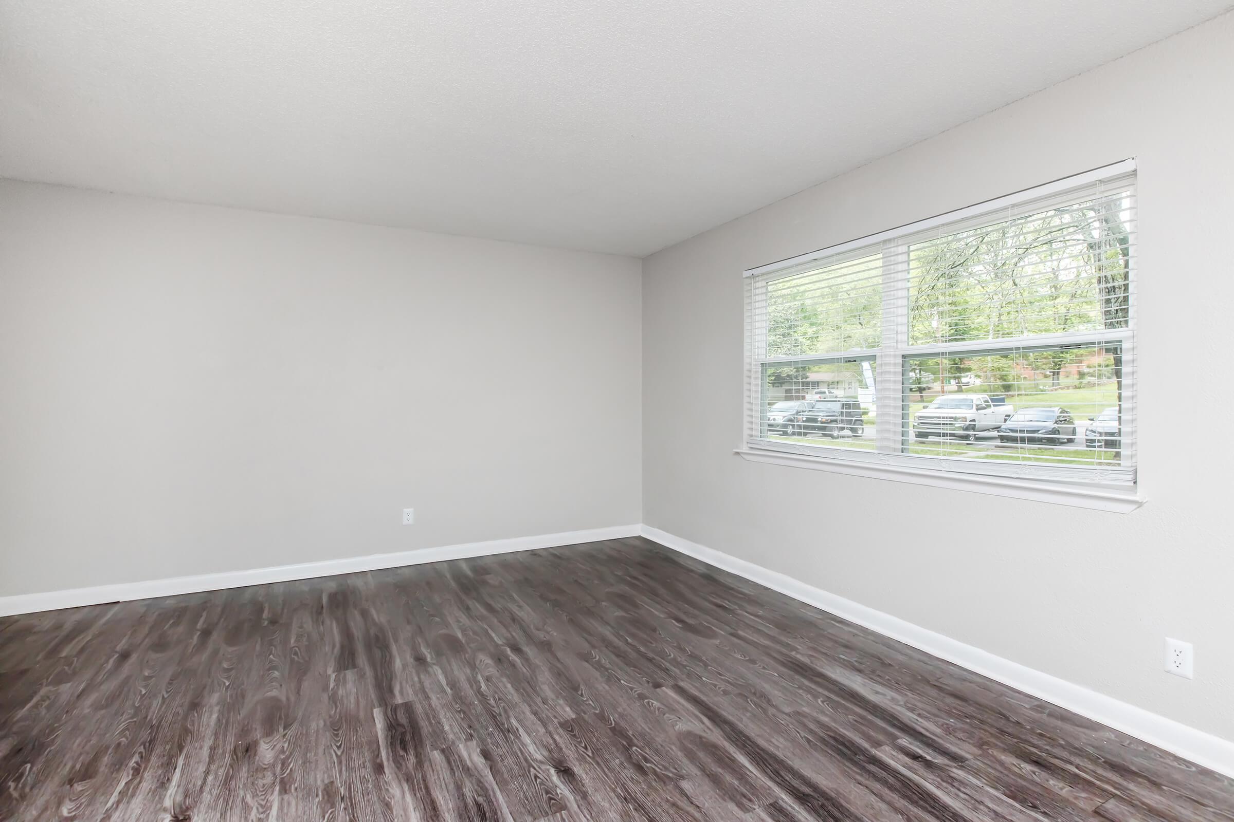 A spacious, empty room with light gray walls and a large window featuring white blinds. The floor is dark-colored, resembling wood. Natural light streams in through the window, illuminating the room, which is free of furniture and decorations.