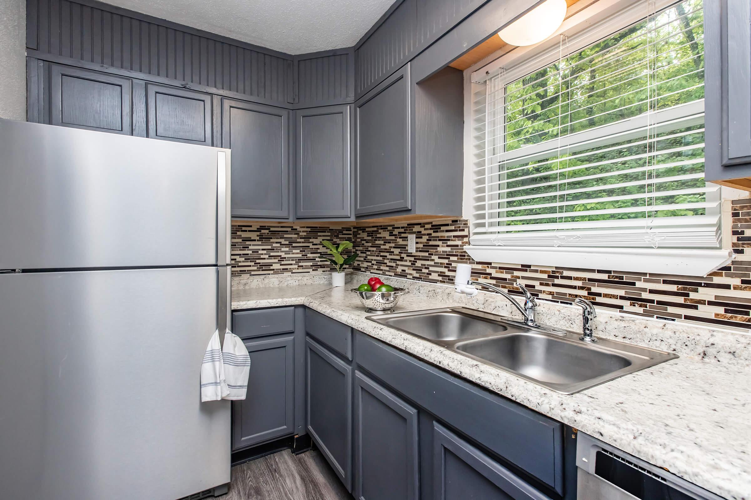 Modern kitchen featuring gray cabinetry, a stainless steel refrigerator, and a granite countertop with double sinks. A bowl of fruit is on the counter, and the window lets in natural light, showing a green outdoor view. The backsplash consists of mosaic tiles for a stylish touch.