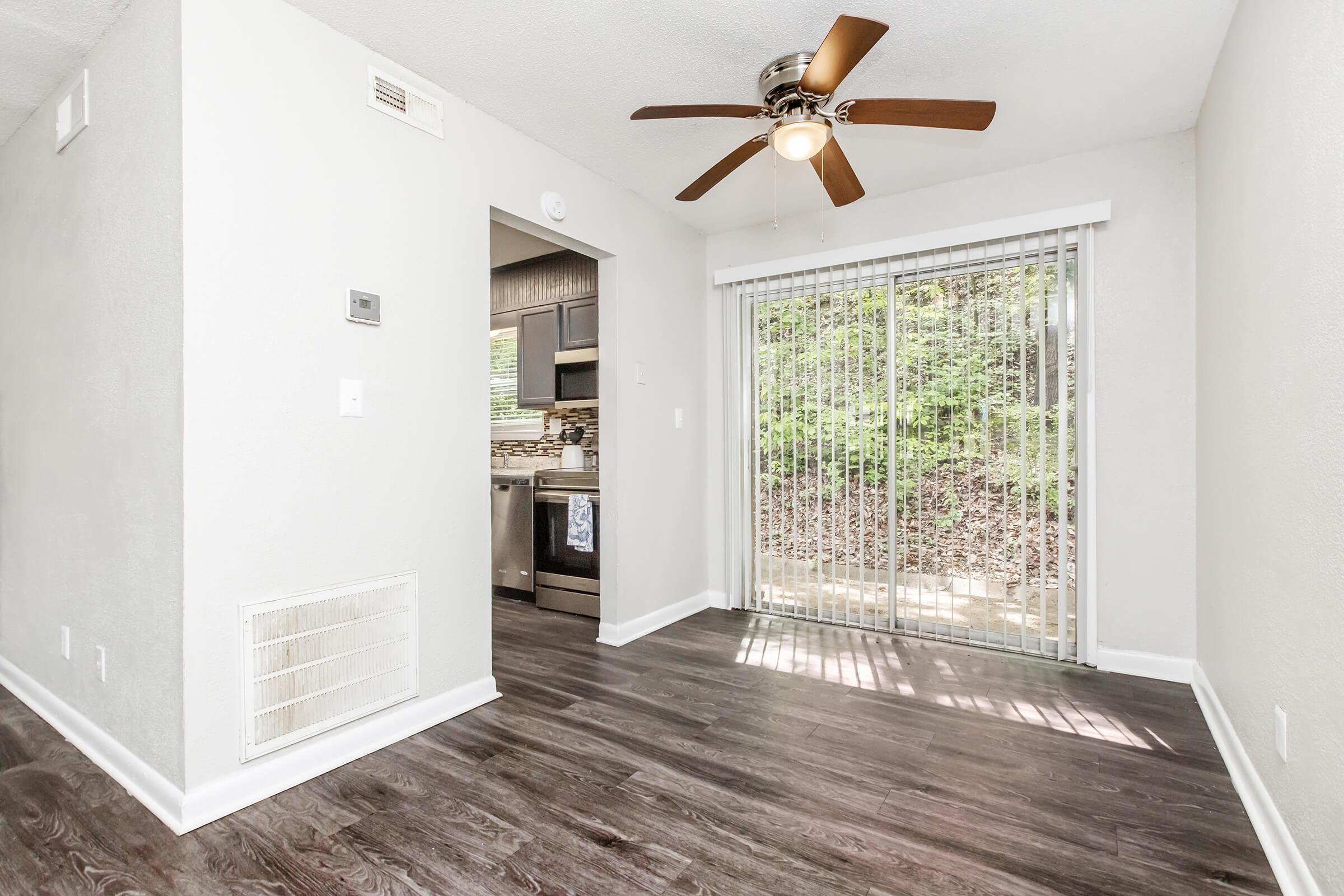 A bright, open living space featuring a ceiling fan, light-colored walls, and dark laminate flooring. A sliding glass door with vertical blinds offers a view of a green outdoor area. The kitchen area is visible in the background, showcasing dark cabinets and appliances.