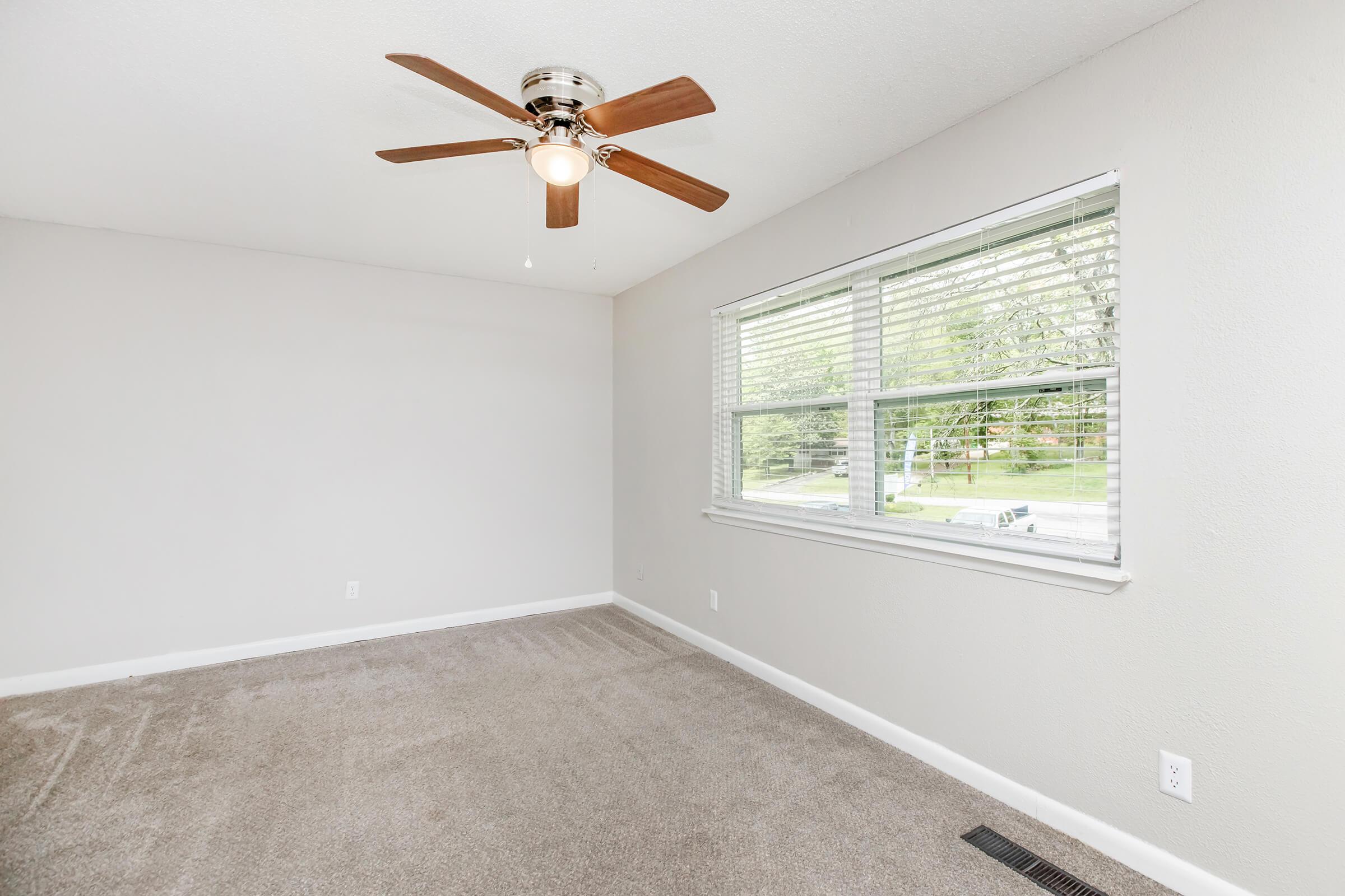 A light and airy room featuring a ceiling fan, carpeted floor, and a large window with blinds. The walls are painted a soft grey, providing a neutral backdrop. Natural light illuminates the space, creating a welcoming atmosphere.