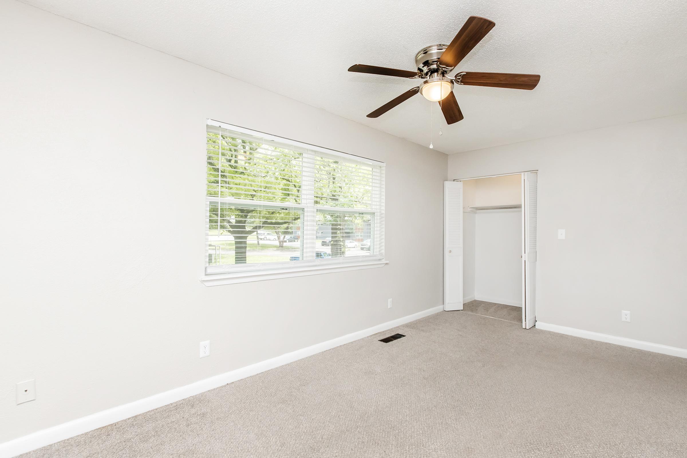 A bright, empty room with light gray walls and tan carpet. A ceiling fan with wooden blades hangs from the ceiling. Large windows provide natural light and show green trees outside. A closet with sliding doors is visible on the right side of the room.