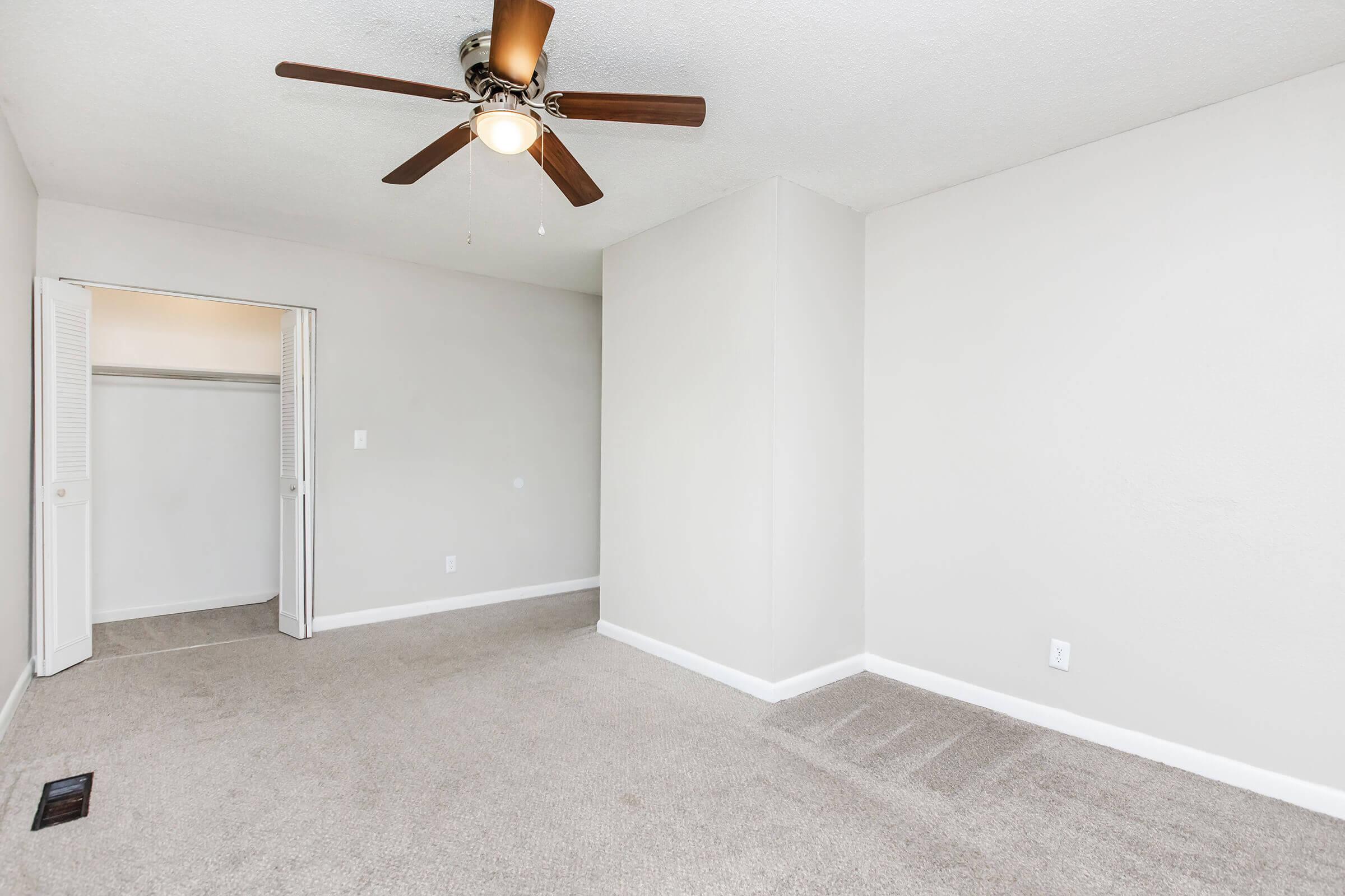 A spacious, empty room featuring light gray walls, a ceiling fan with wooden blades, and plush beige carpet. There's an open closet with sliding doors to the left, and the room is well-lit with natural light coming from an unseen window.