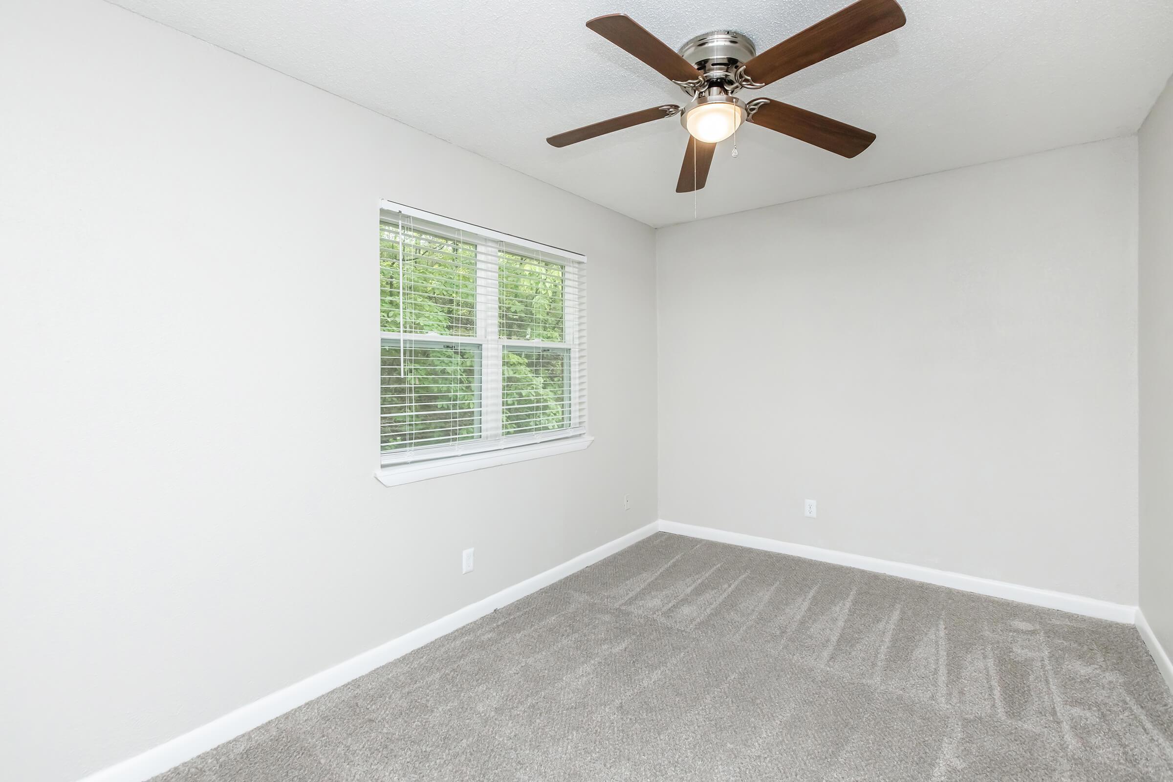 A vacant bedroom with light grey walls and a light grey carpet. It features a ceiling fan with a light fixture and a window with white blinds, allowing natural light to enter the room. The space is empty, creating a clean and minimalistic atmosphere.