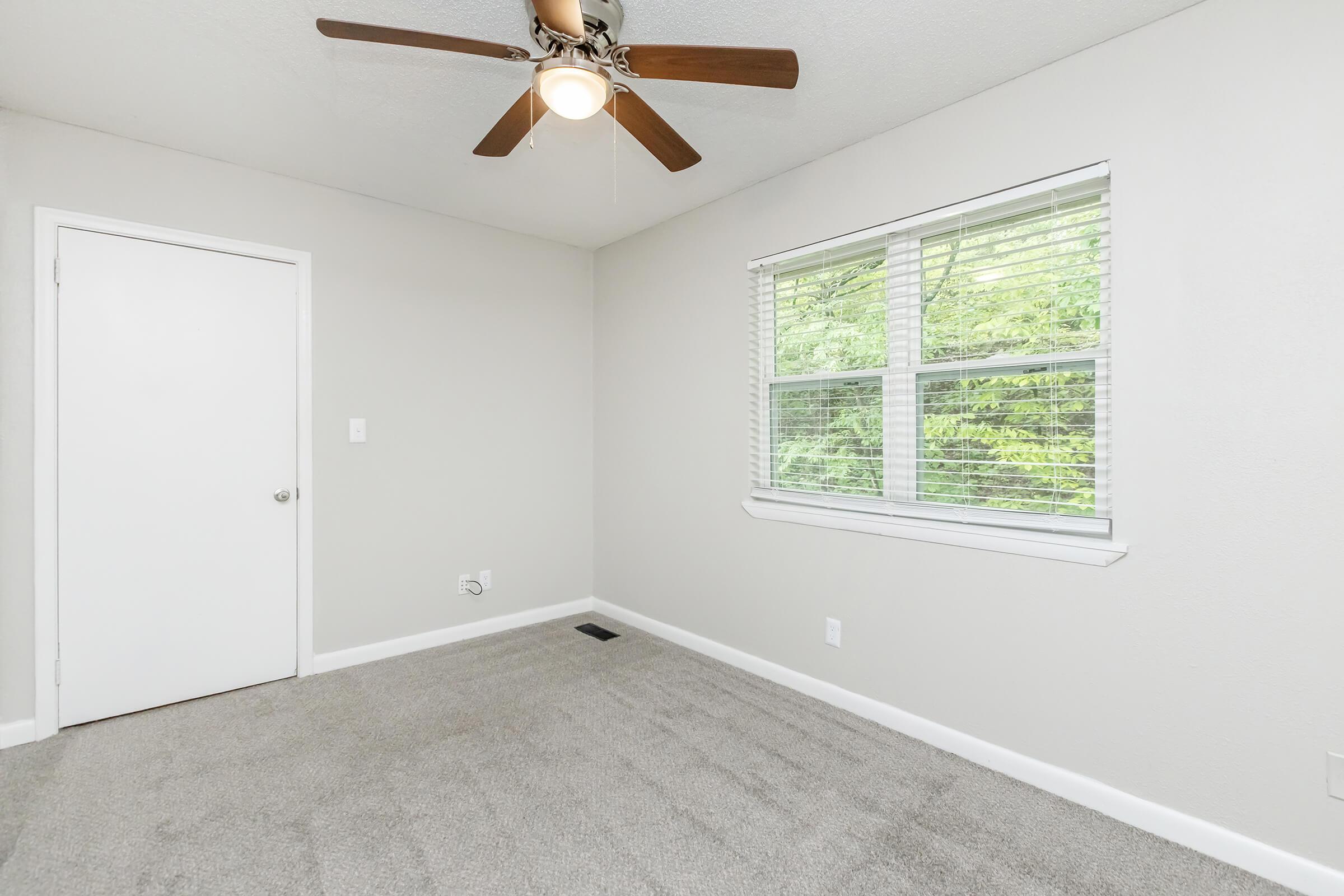 A clean, empty room featuring light gray walls and new carpet. A ceiling fan hangs from the ceiling, and there's a window with blinds, allowing natural light to enter. A single white door is visible, and the overall atmosphere is bright and inviting.
