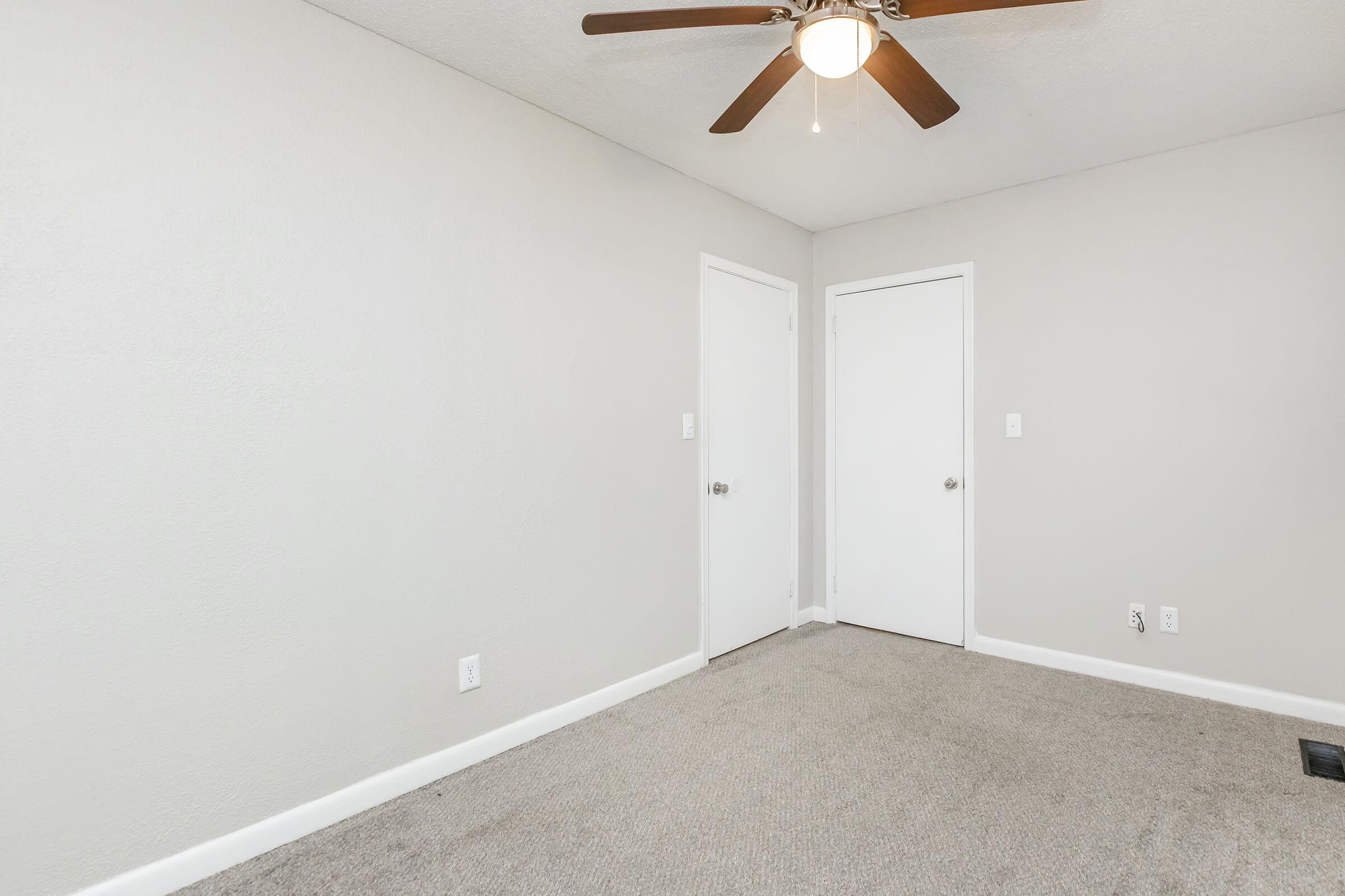 A vacant room with light gray walls, a ceiling fan with wooden blades, and beige carpet. There are two white doors, one leading to a closet and the other to a space outside the room. The lighting is bright, and the space appears clean and uncluttered.