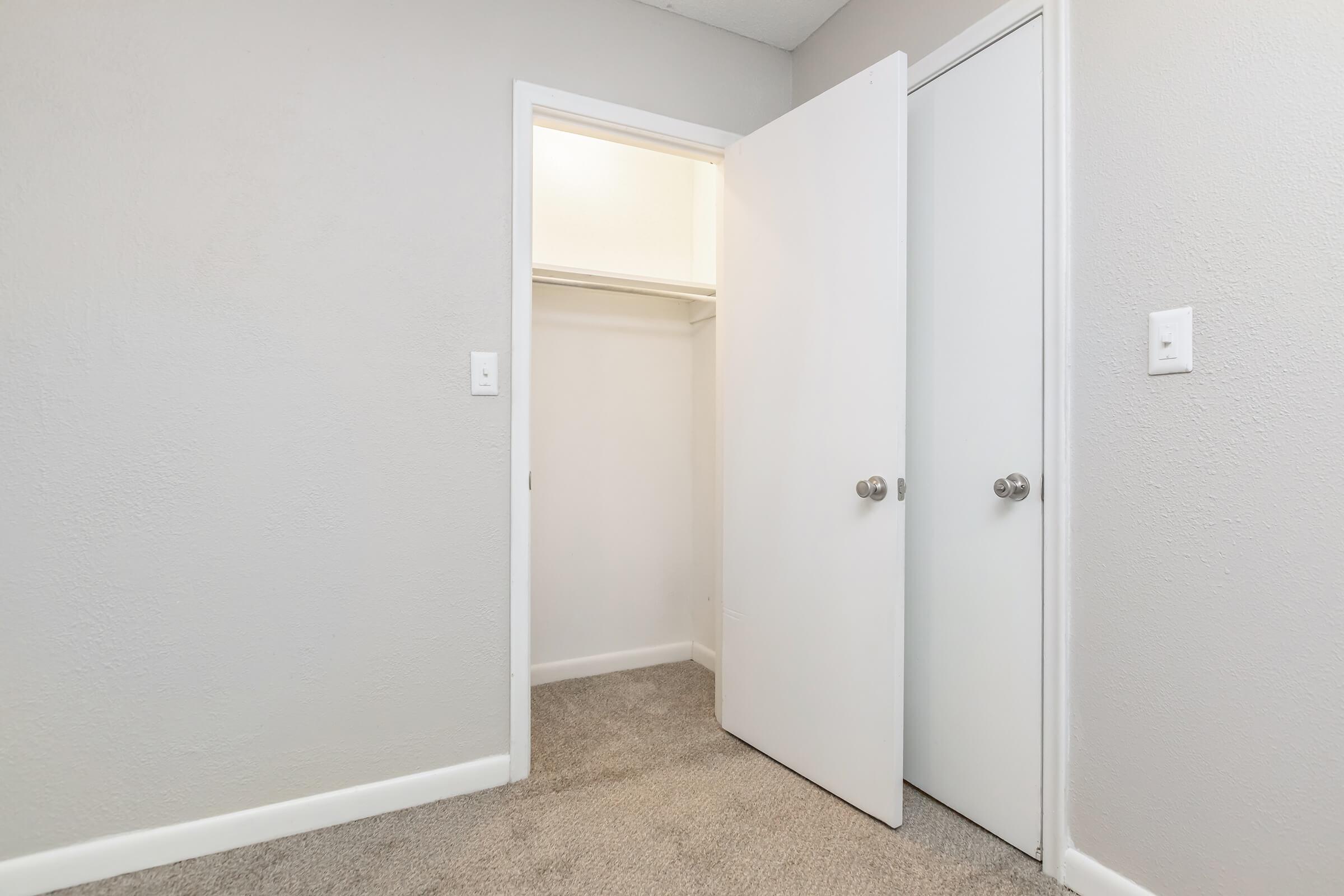 A small room featuring a light gray wall with a beige carpeted floor. Two white doors are partially open; one leads to a closet, while the other appears to be a standard door. The lighting is soft, creating a clean and minimalistic atmosphere.