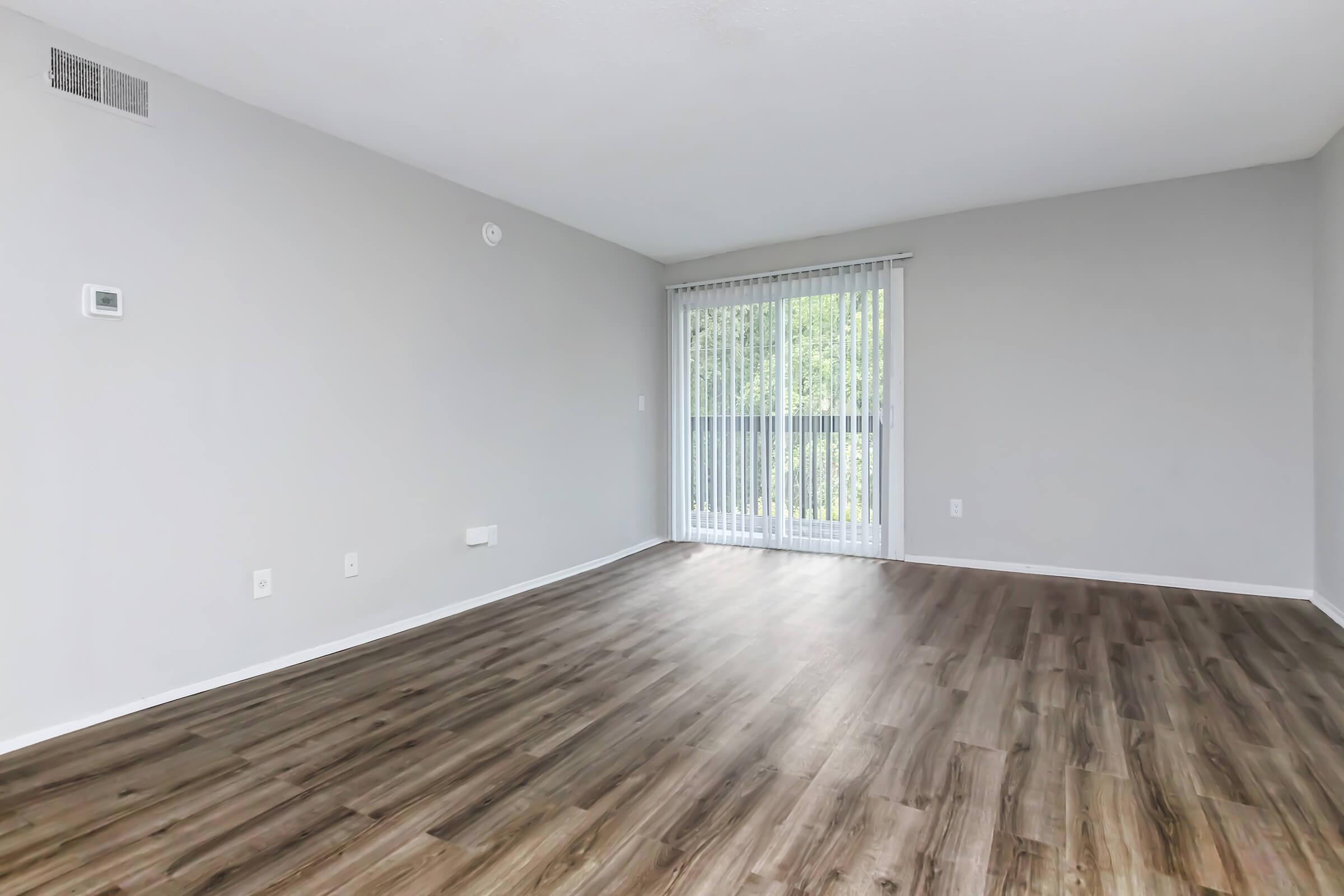 Spacious interior of a modern living room with light gray walls and hardwood-style flooring. Large sliding glass door with vertical blinds allows natural light, leading to a balcony. A thermostat is mounted on the wall, and the space is empty, providing room for furniture and decor.