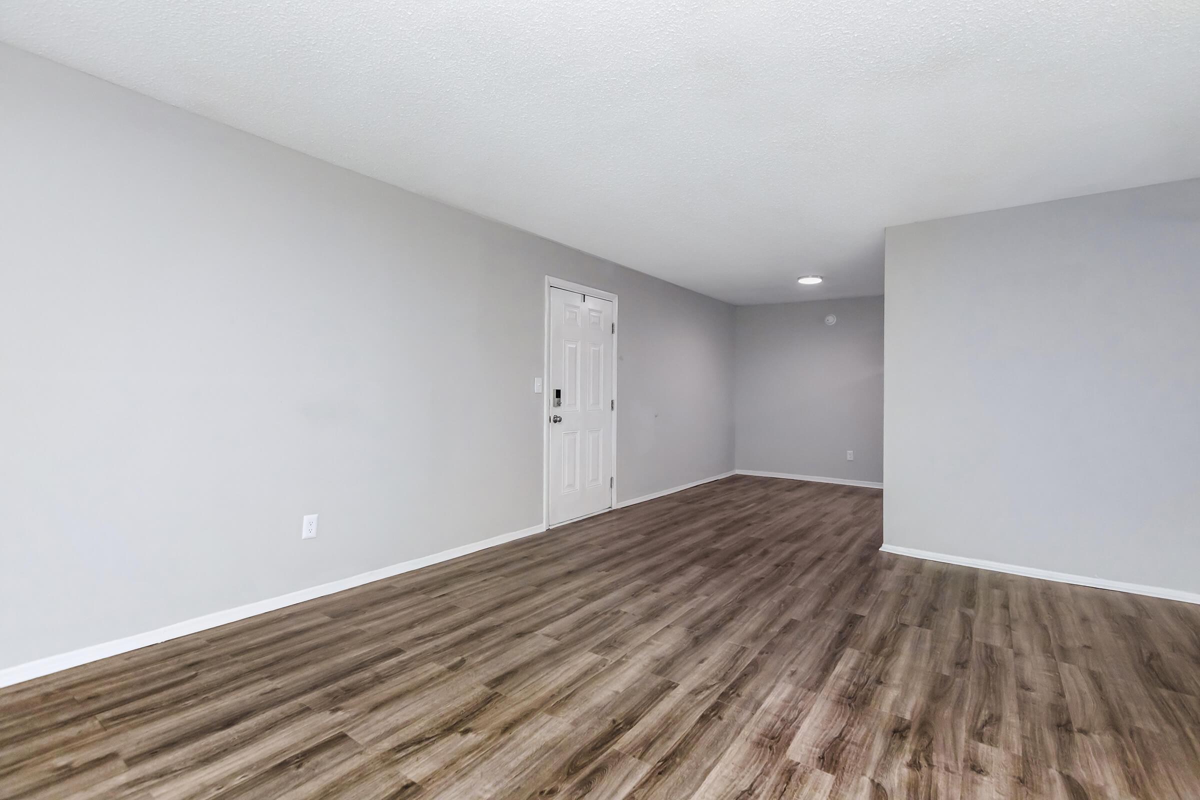 A spacious, empty room featuring light gray walls, new wooden flooring, and a single white door. The area is well-lit with a ceiling light and has no furniture, creating a clean and open atmosphere suitable for various layouts or purposes.