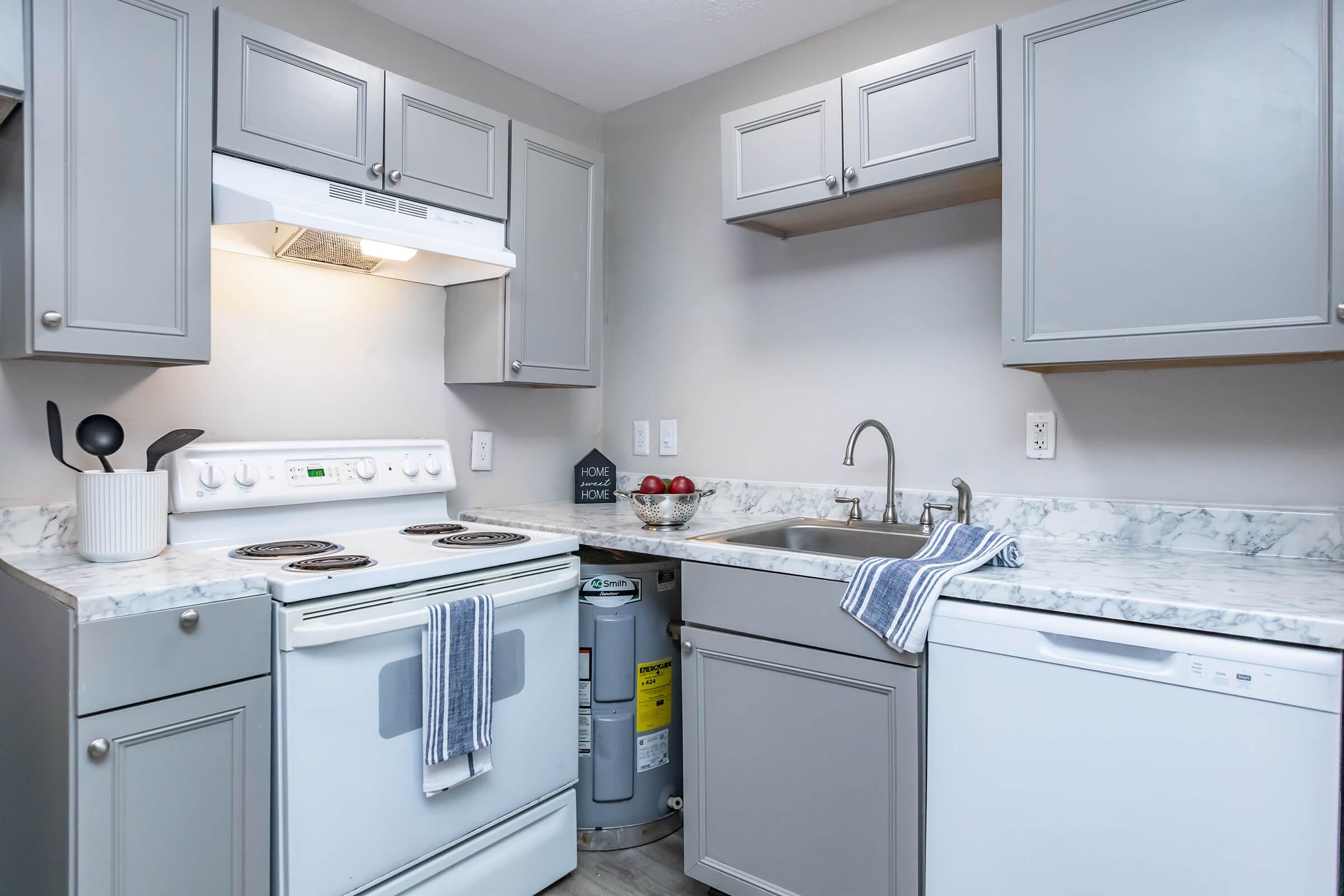 A modern kitchen featuring gray cabinets, a white stove, and a dishwasher. The countertop has a marble finish, with a sink and decorative items. A small water heater is visible beneath the sink. The space is well-lit and organized, creating a clean and functional environment.