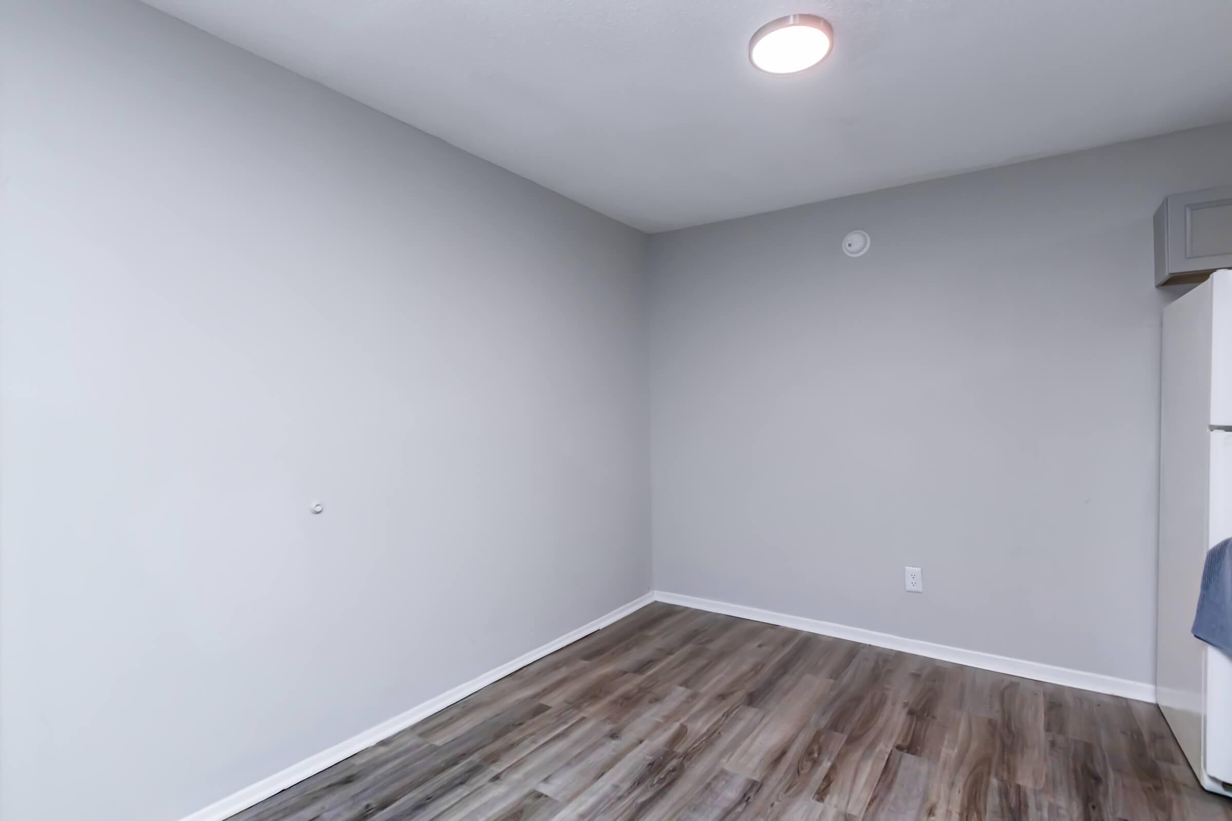 Empty room with light gray walls and a wooden floor. A ceiling light fixture is visible, and there is a white refrigerator in the corner. The room is unadorned, creating a minimalist appearance.