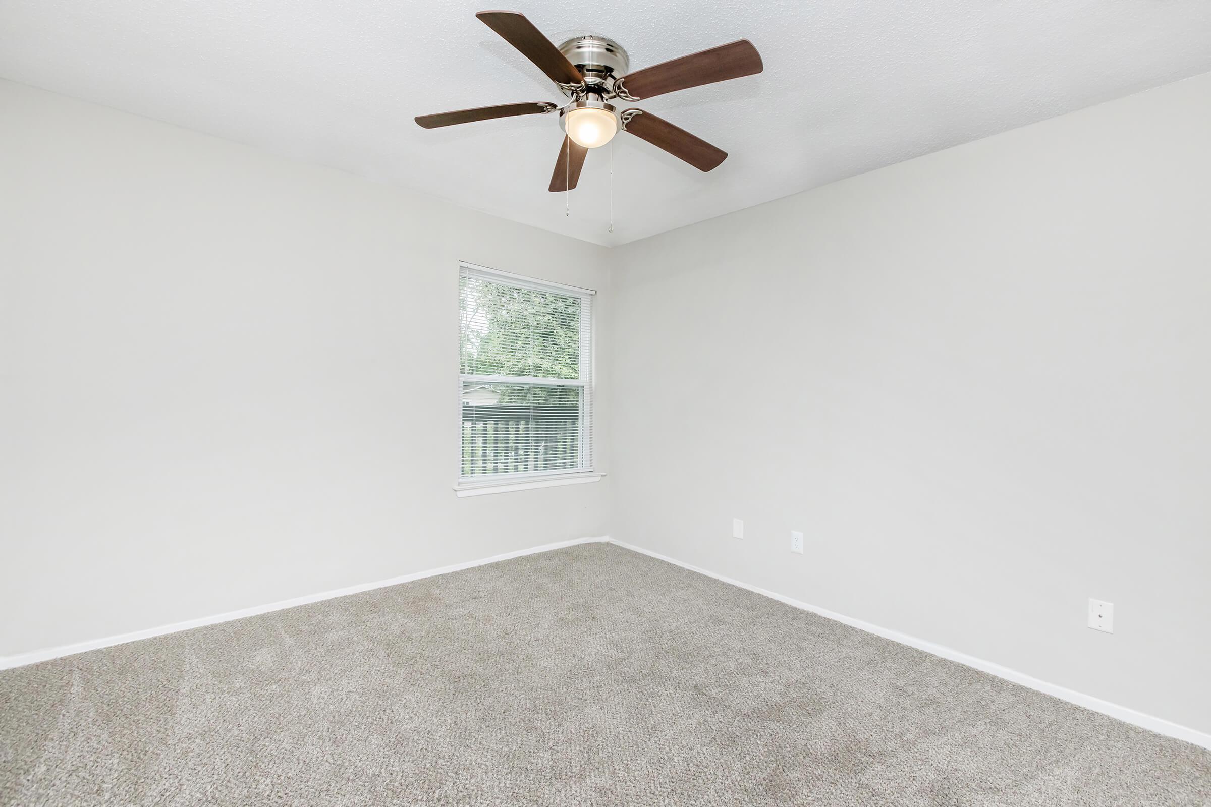 A vacant room featuring light gray walls, beige carpet flooring, and a ceiling fan with wooden blades. There is a window allowing natural light, which enhances the spacious feel of the area. The room has no furniture, creating a blank canvas for potential occupants.