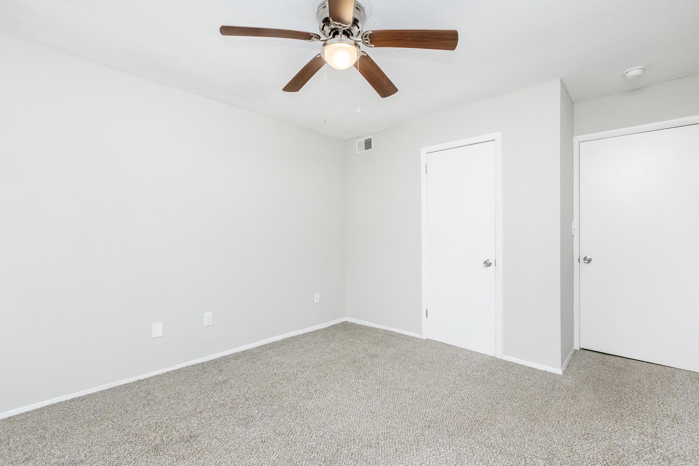 A vacant room featuring light gray walls and a light-colored carpet. A ceiling fan with wooden blades is mounted in the center. There are two doors; one is closed, likely leading to a closet, and the other is a standard door. Natural light can be seen entering the room.