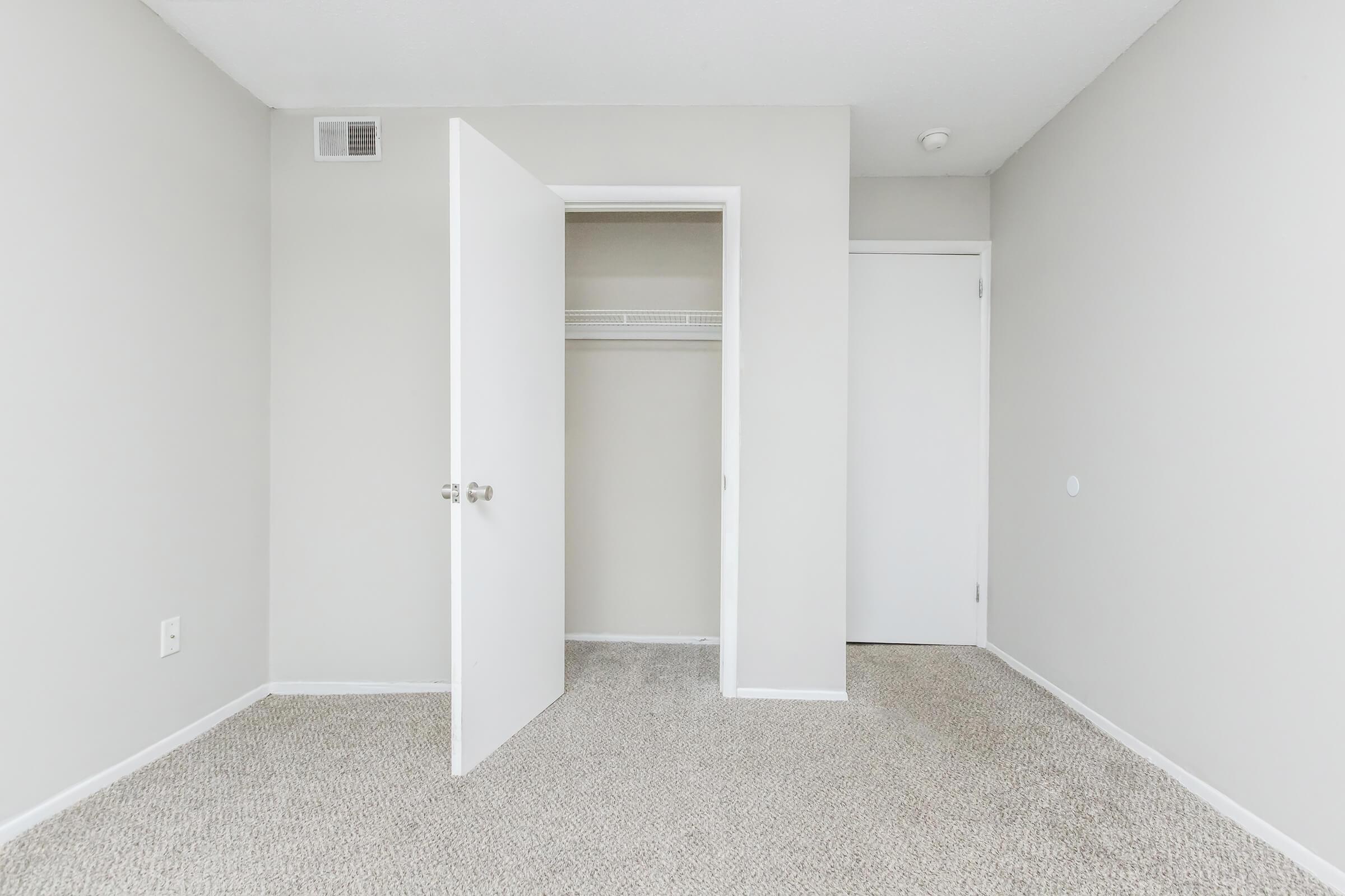 Empty room with light gray walls and beige carpet. There are two doors, one leading to a closet and the other to an adjacent space. The room has no furnishings and features a small wall vent. Natural light brightens the space, emphasizing its simplicity and cleanliness.
