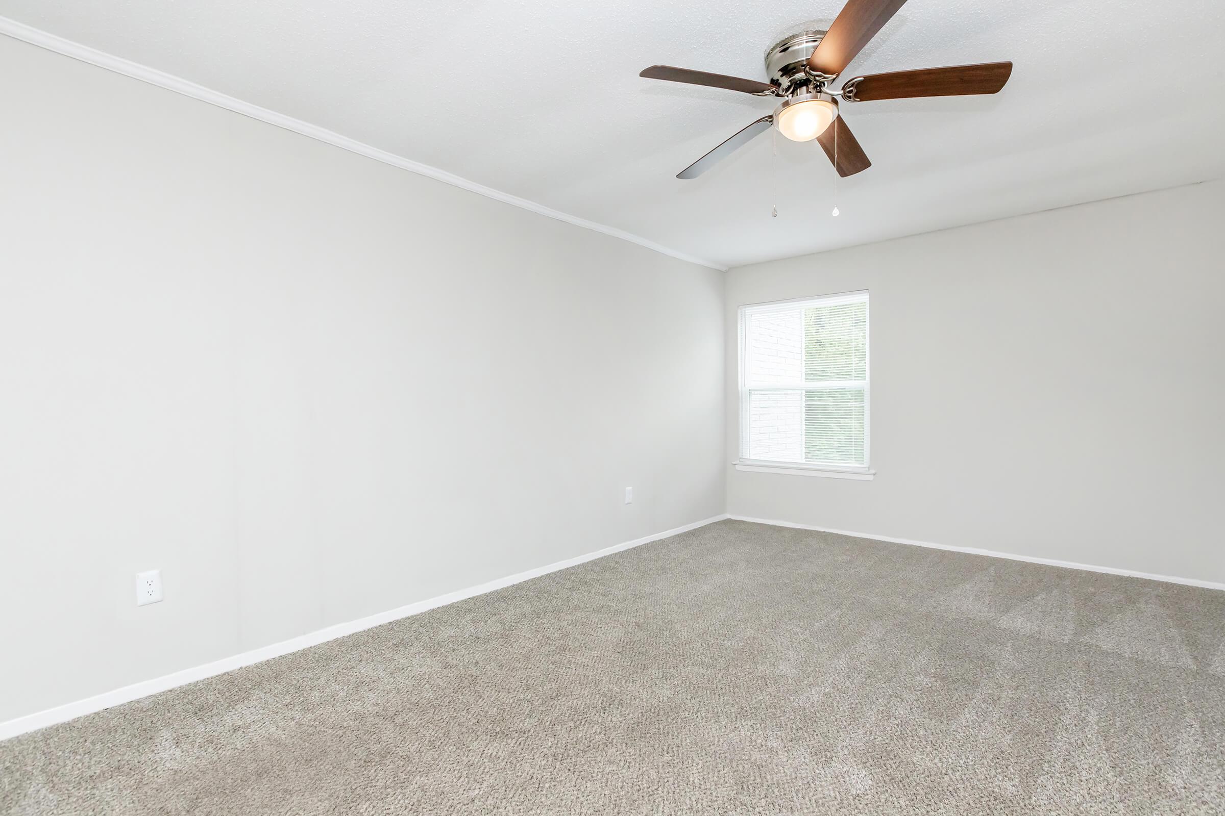 Empty room with light gray walls and carpeted flooring. A ceiling fan with wooden blades is mounted on the ceiling. A single window allows natural light to enter the space. The room has a clean, minimalistic design, suitable for various uses.