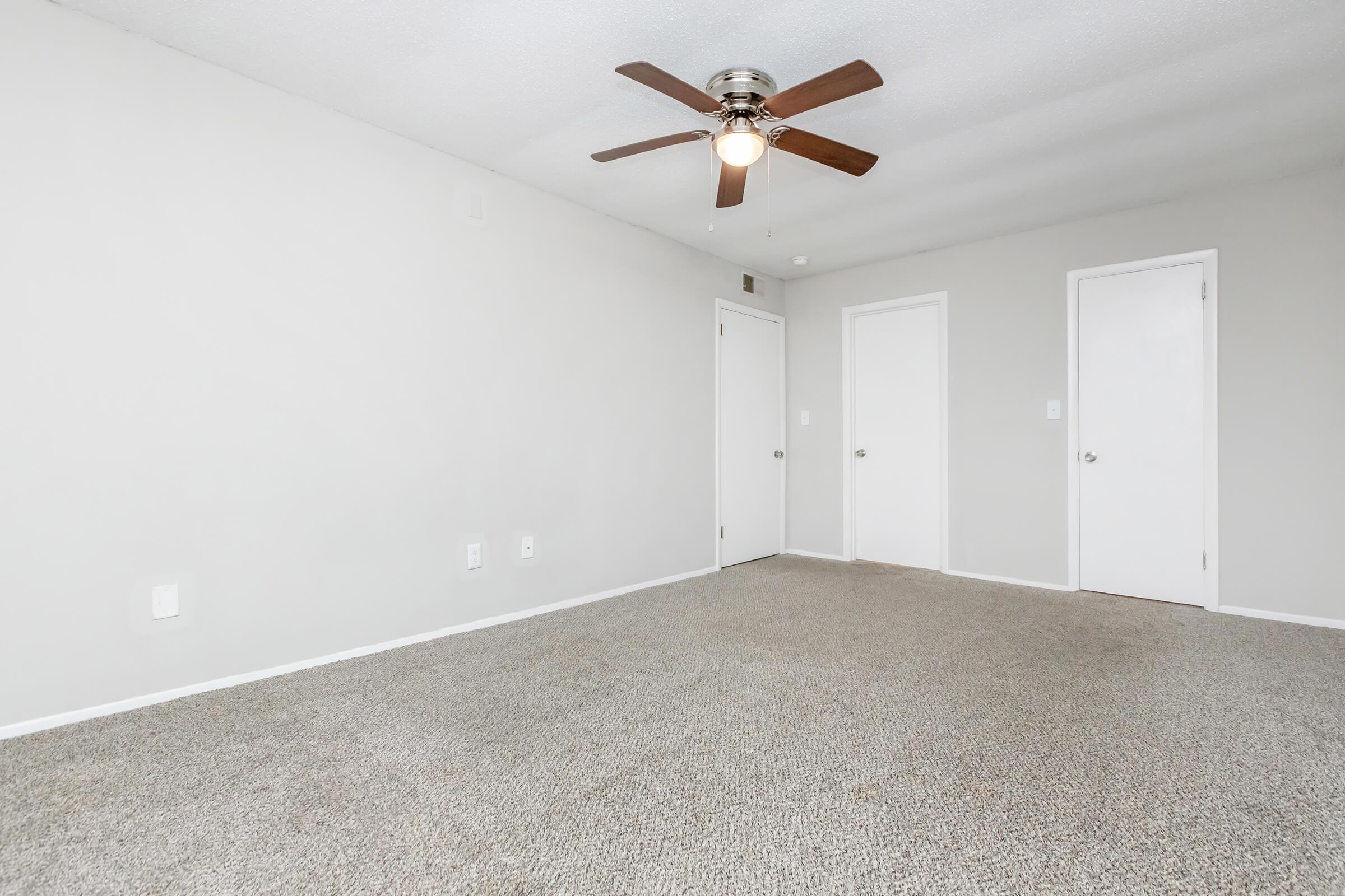 A spacious, lightly colored room with beige carpet flooring. It features a ceiling fan with a light, two white doors on the right, and plain walls. The overall atmosphere is clean and minimalistic, ideal for various uses like a bedroom or office.
