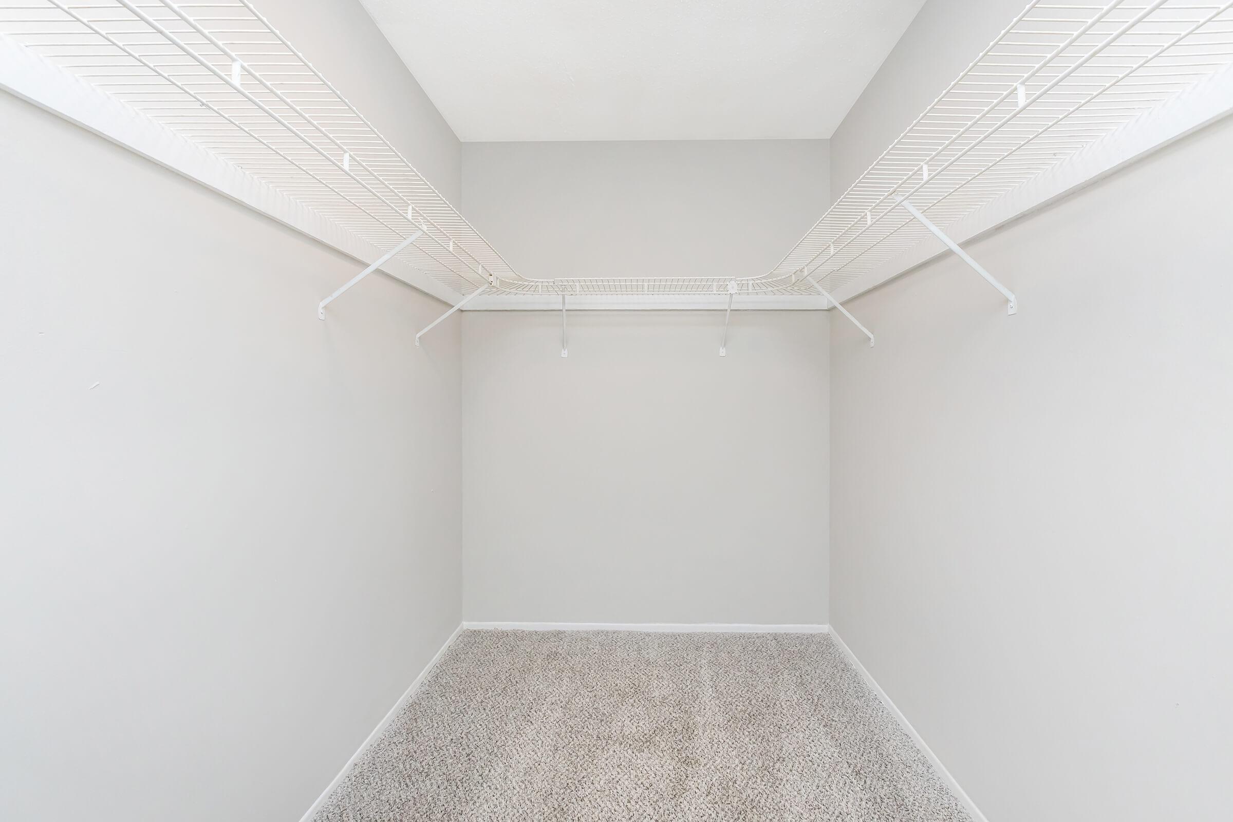 Empty closet with gray walls and a carpeted floor. The closet features a wire shelf along the top, with no items stored inside, creating a spacious and minimalist appearance.