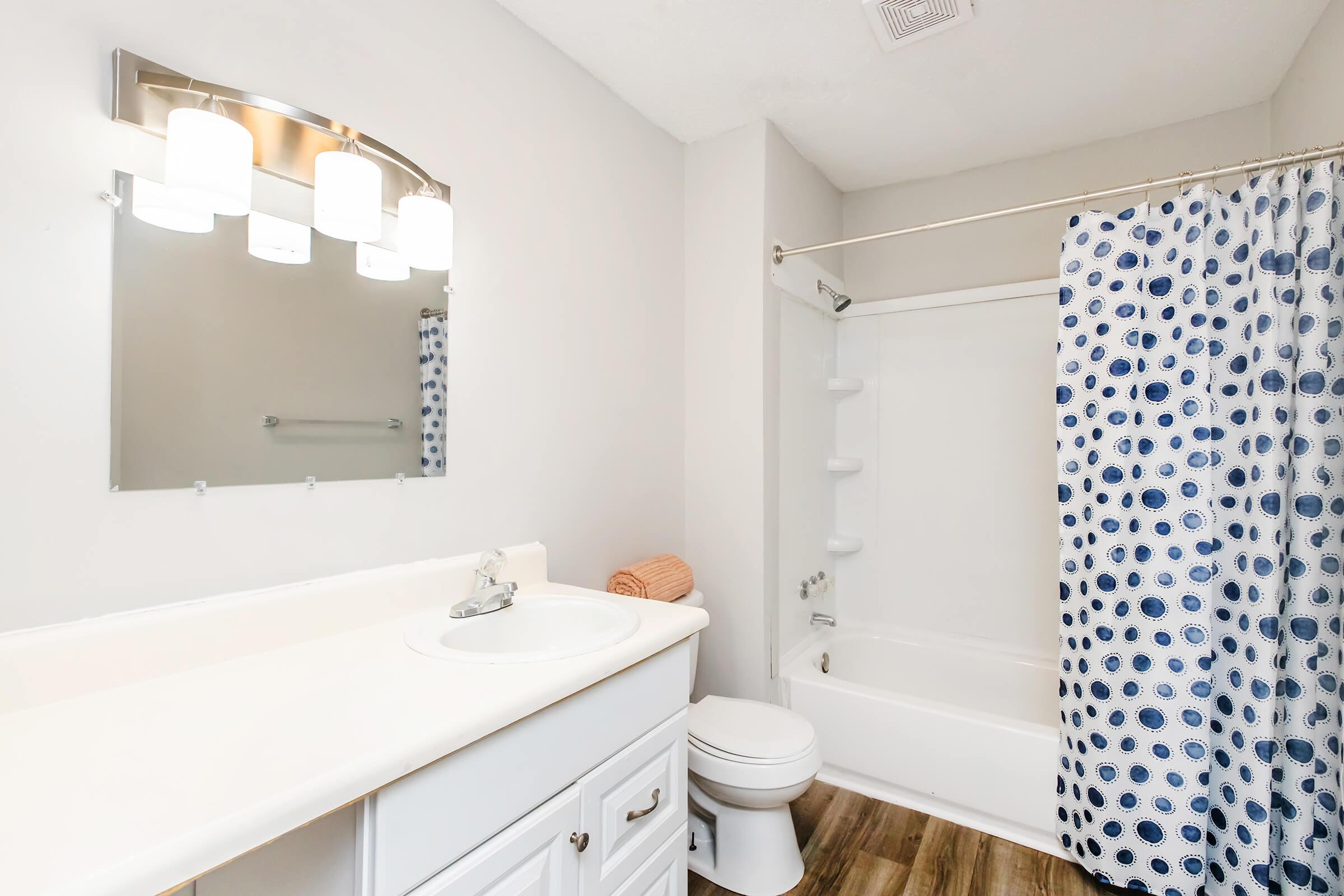 A clean bathroom featuring a white sink with a silver faucet, a large mirror with three light fixtures above, a shower/tub combination with a blue and white polka dot curtain, and a modern toilet. The floor is wood-like, and there is an orange towel rolled up on the countertop.