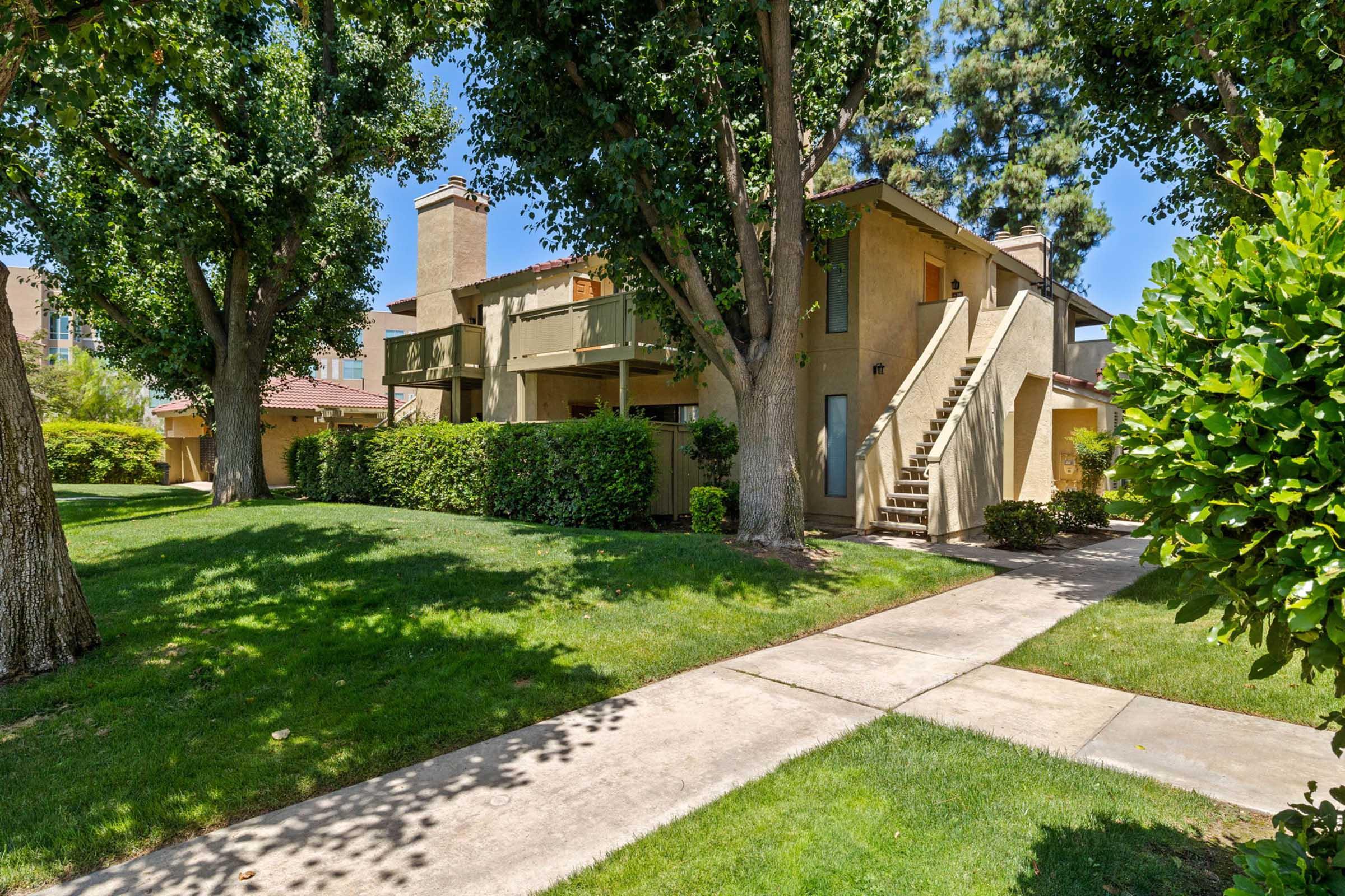 A well-maintained apartment building featuring two stories with balconies, surrounded by lush green grass and trees. A concrete pathway leads through the vibrant landscaping, creating a welcoming outdoor space. The sky is clear and blue, enhancing the pleasant atmosphere.