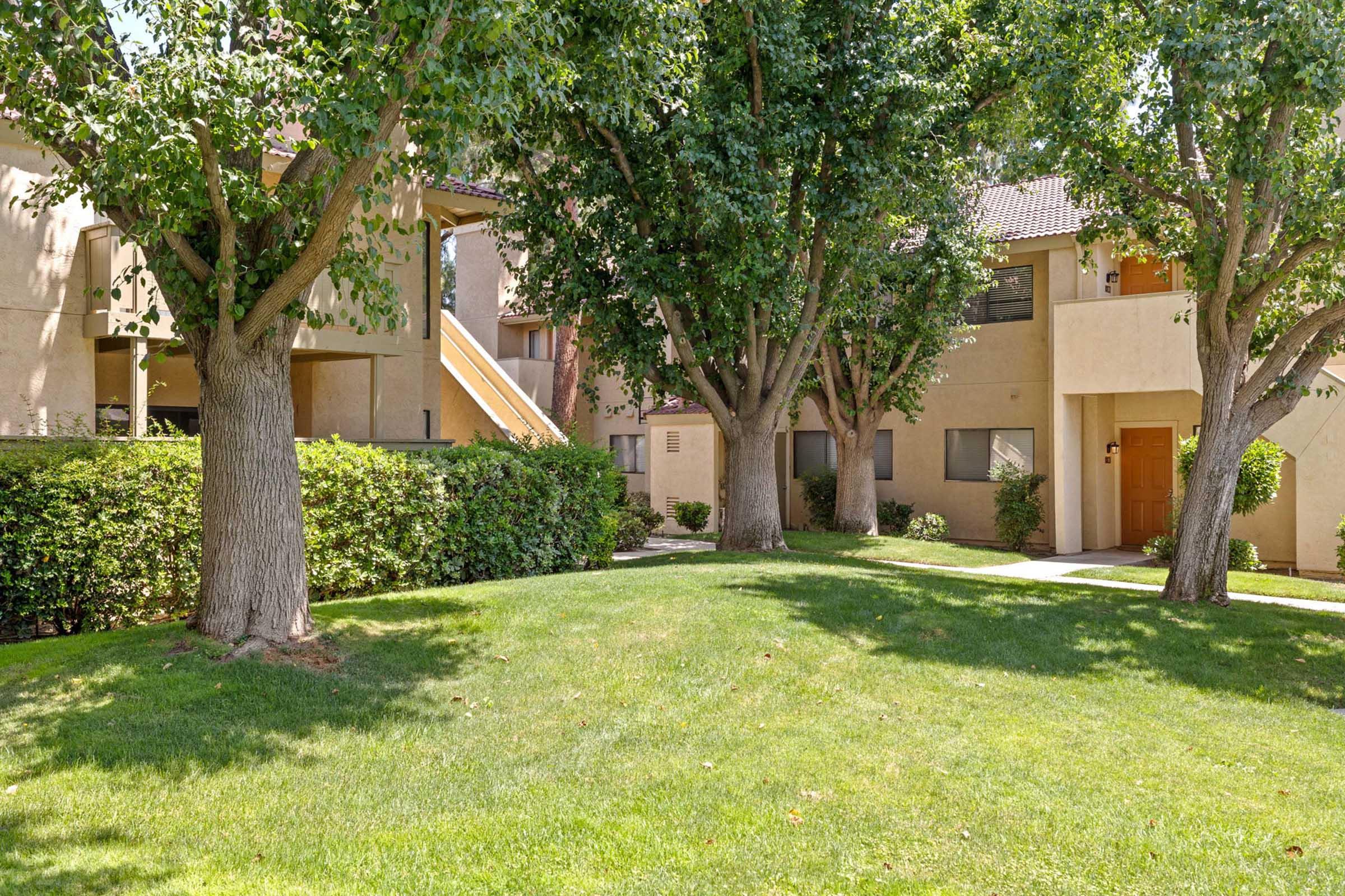 A landscaped area featuring two buildings with light-colored exteriors, surrounded by grassy lawns and several leafy trees. The scene is bright and inviting, showcasing a well-maintained residential environment.