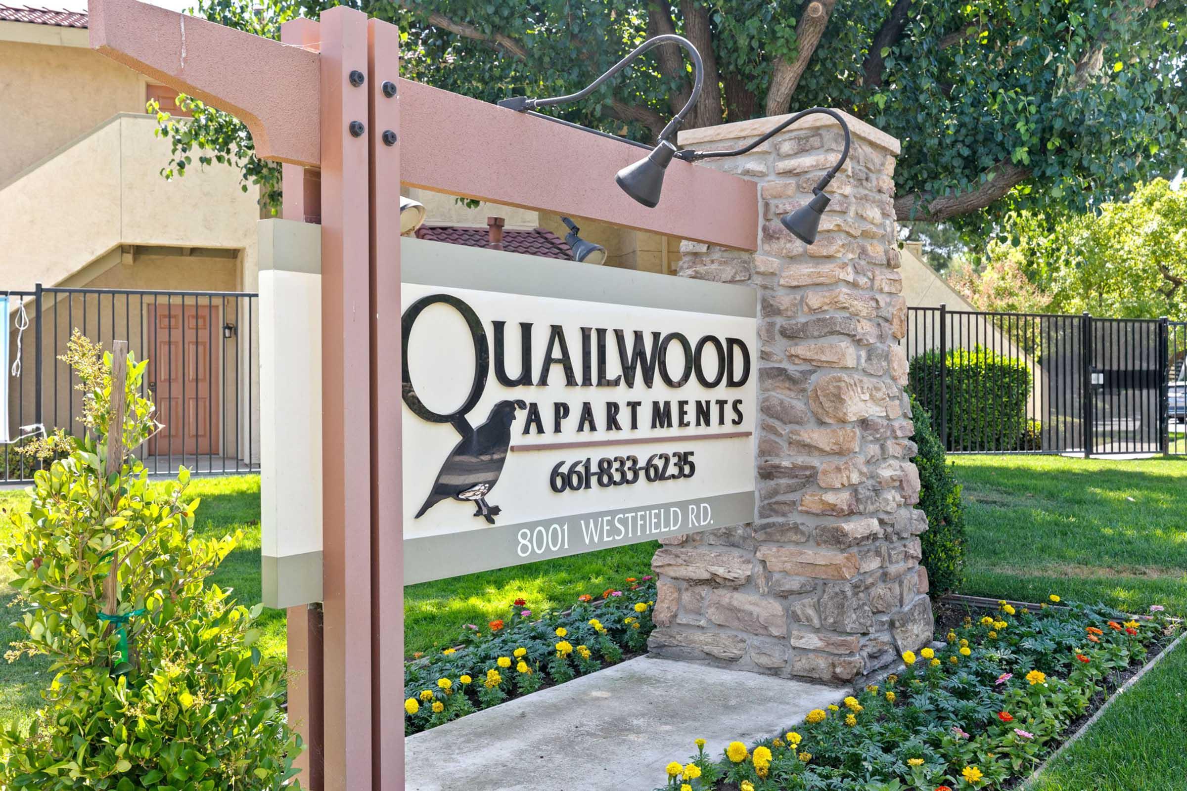 Sign for Quailwood Apartments located at 8001 Westfield Rd, featuring a decorative design with a bird silhouette. Surrounding the sign are landscaped plants and flowers, with a gated entrance visible in the background.