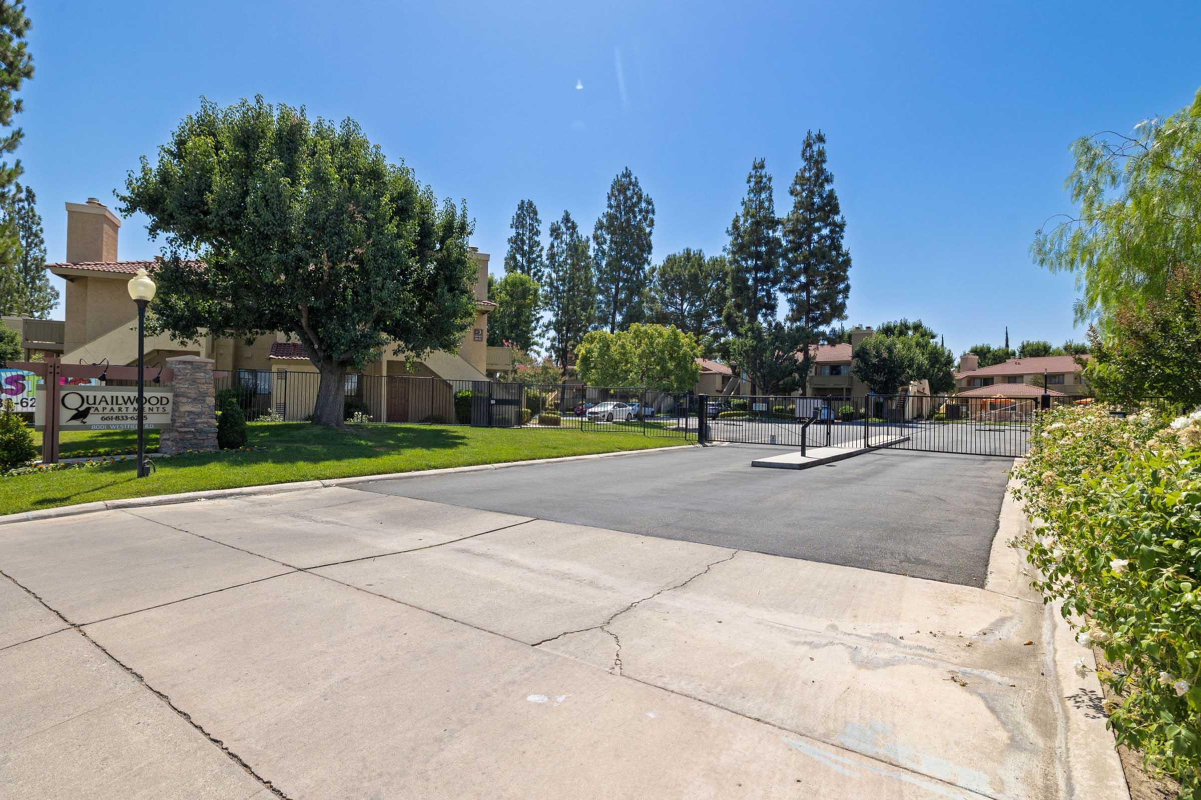 A view of a residential complex entrance featuring a gated entry. There are trees and landscaped greenery lining the sides of the driveway, with several buildings visible in the background under a clear blue sky.