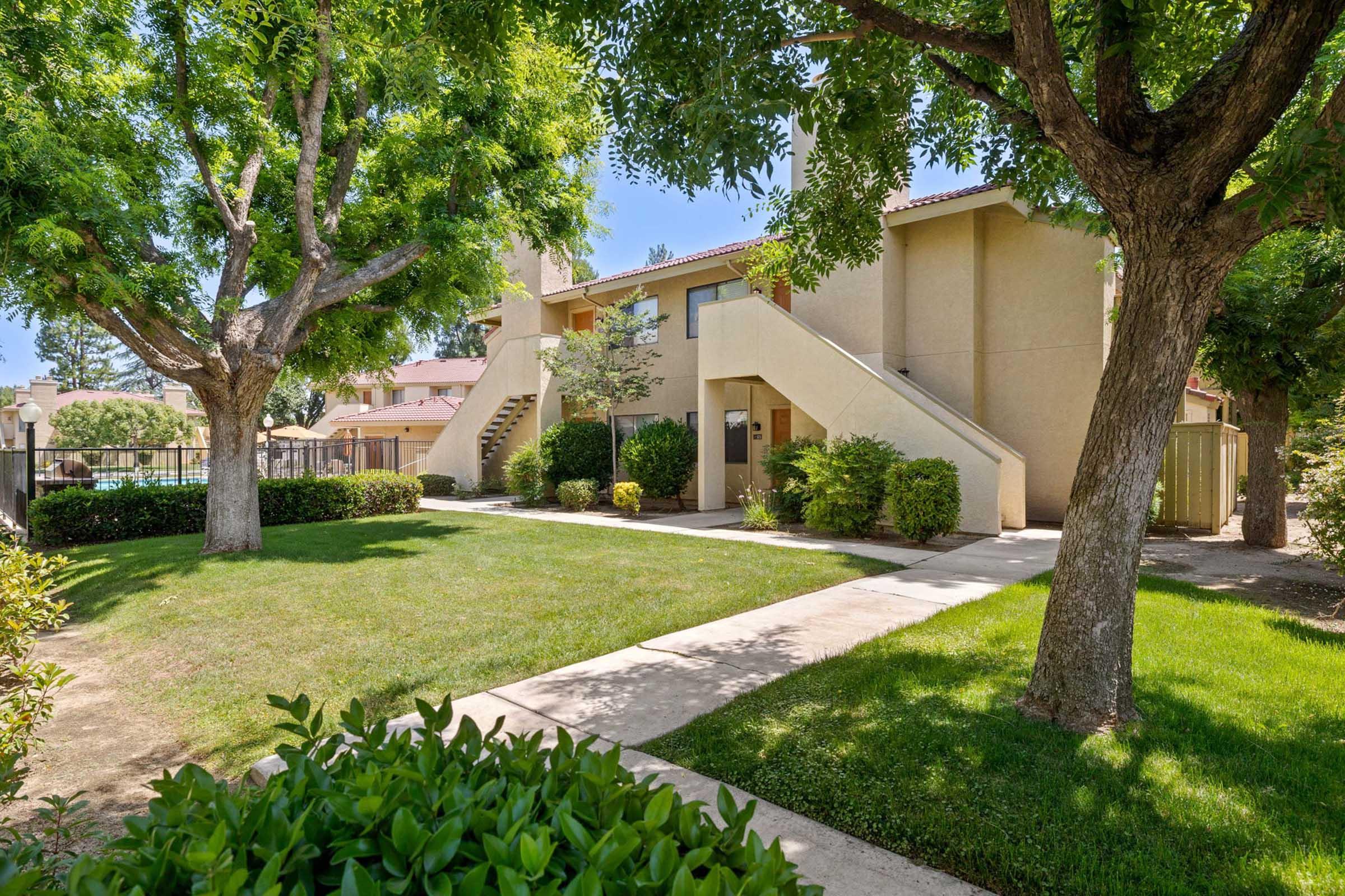 Two-story apartment building surrounded by well-maintained grassy areas and trees. There are stairs leading to the second floor, and a pathway runs through the greenery. The scene is bright and sunny, suggesting a pleasant outdoor environment near the building.