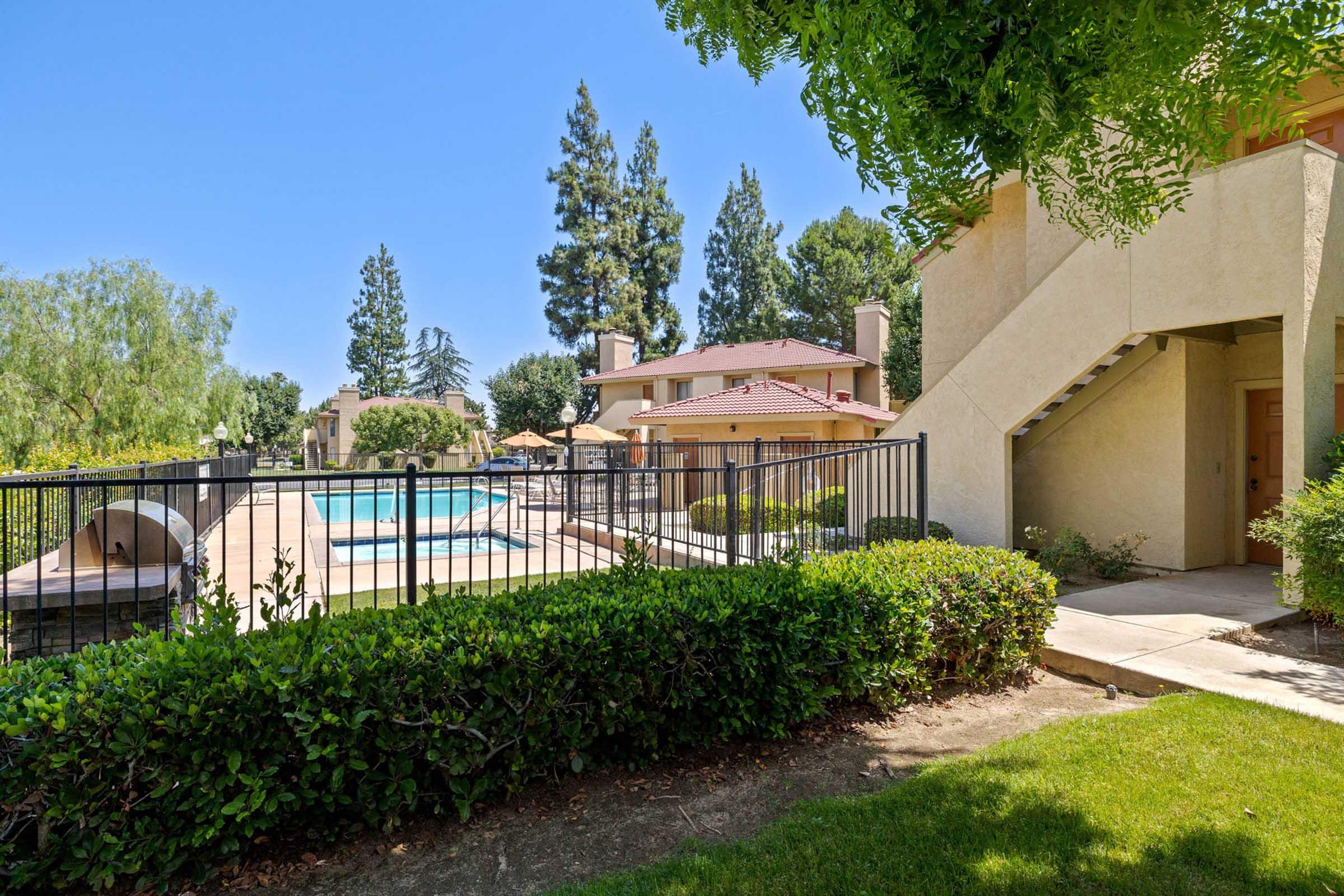 View of a well-maintained outdoor area featuring a pool surrounded by lounge chairs and umbrellas, framed by lush greenery and tall trees. A two-story building with a staircase is located nearby, offering a peaceful atmosphere for relaxation and leisure.