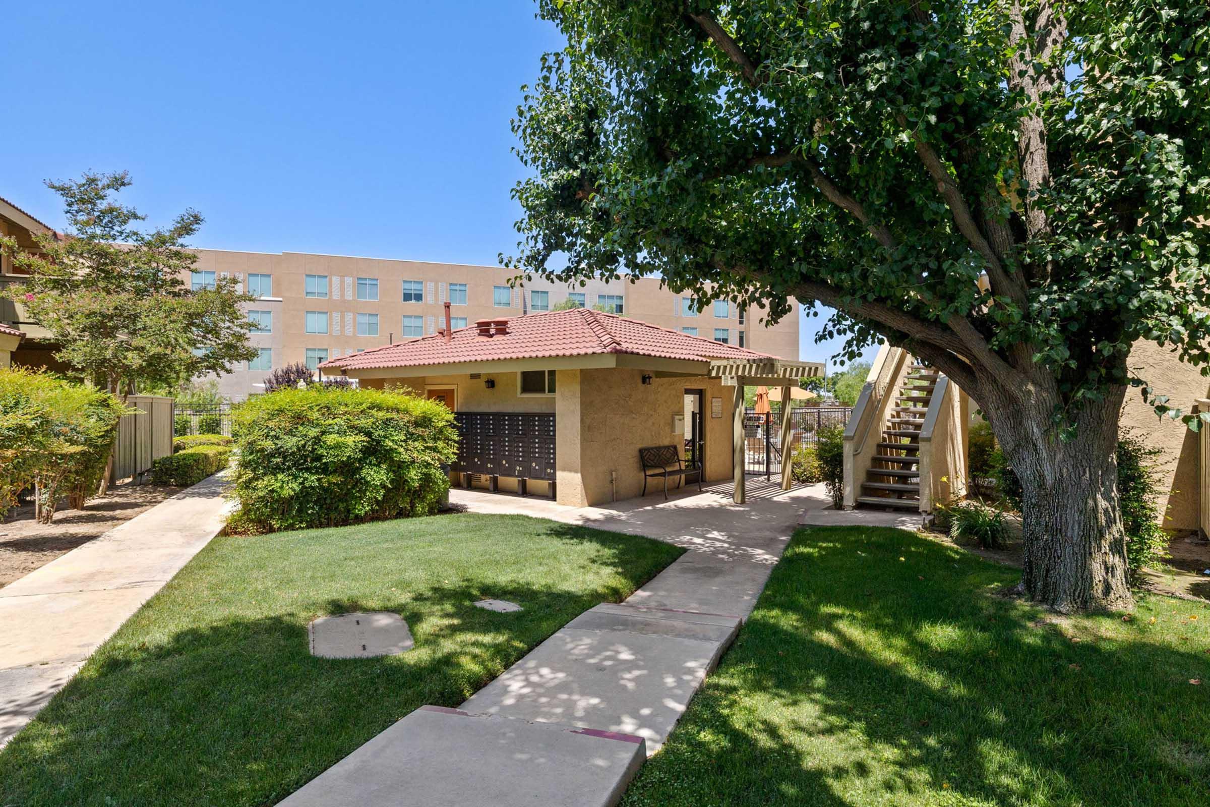 A well-maintained courtyard featuring a path leading to a building with mailboxes and stairs. Lush green grass and shrubs surround the area, with a large tree providing shade. In the background, there is a multi-story building visible against a clear blue sky.