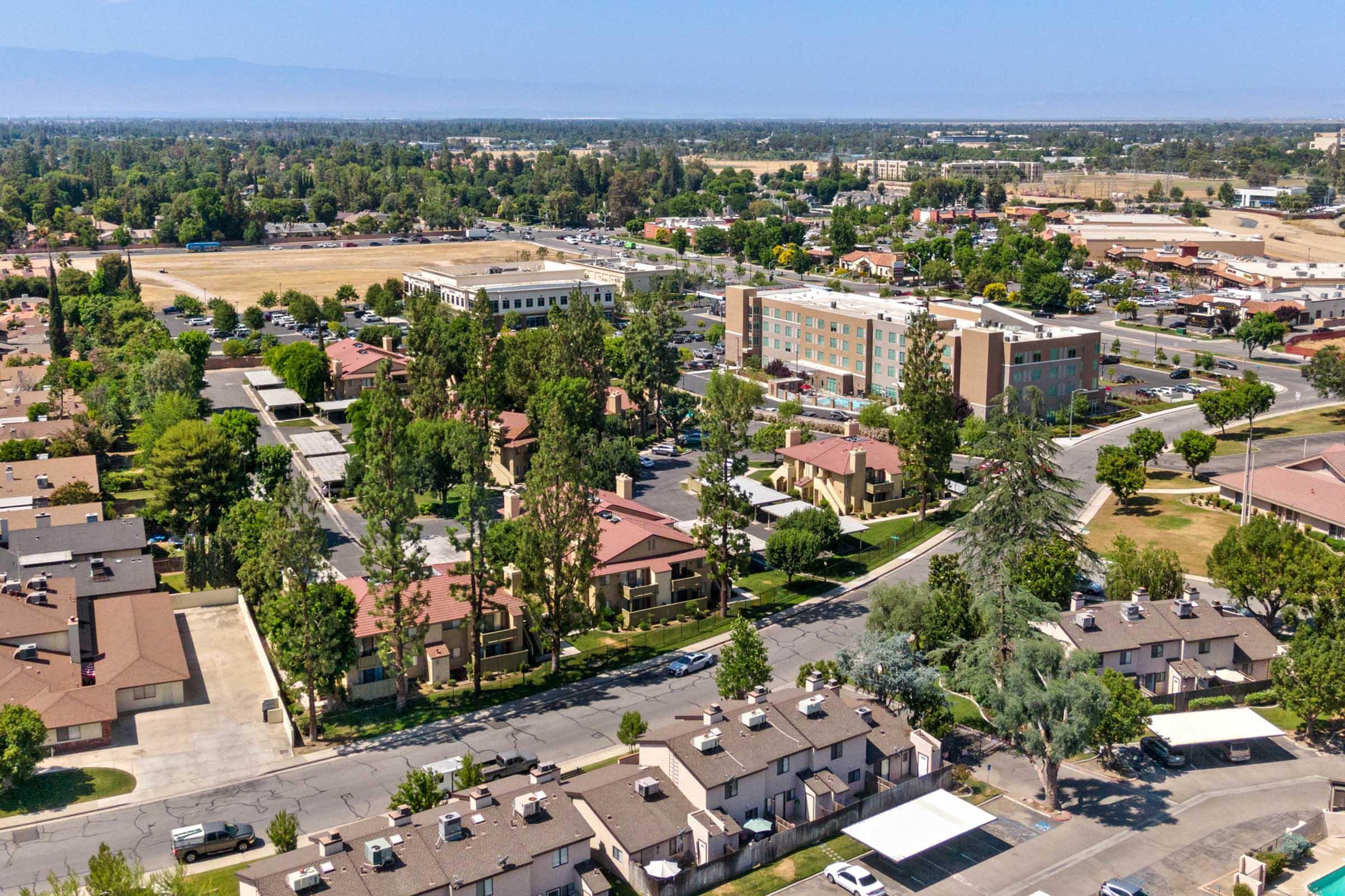 Aerial view of a suburban neighborhood featuring low-rise residential buildings with red roofs, green trees, and well-maintained streets. In the background, there are commercial buildings and open fields under a clear blue sky. The area appears lush and developed, typical of a small city landscape.