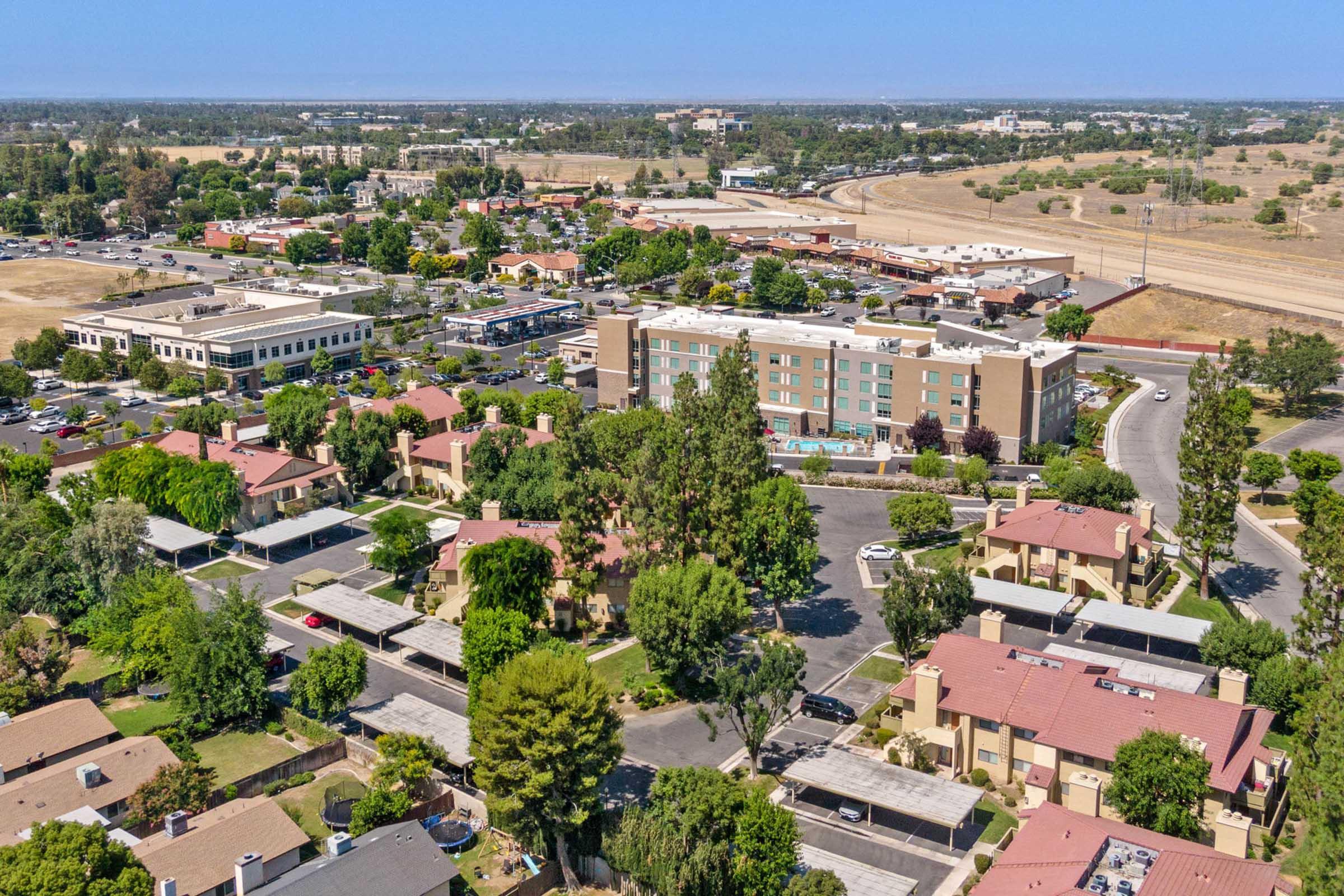 Aerial view of a suburban area featuring several buildings, including residential complexes with red roofs and a hotel. The scene includes a shopping plaza, green trees, and expansive parking lots, with roads visible in the background leading to open fields.