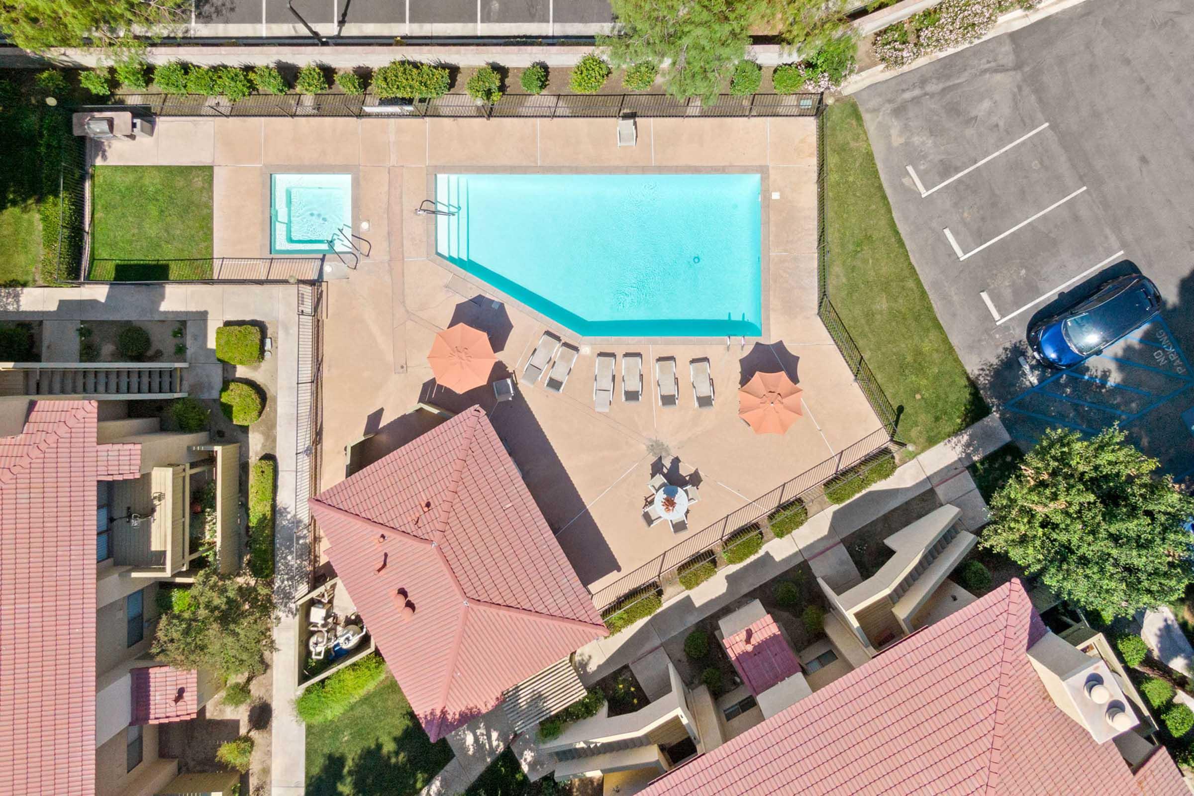 Aerial view of a residential area featuring a swimming pool surrounded by lounge chairs and umbrellas. Nearby are landscaped green spaces and apartment buildings with red roofs. An adjacent parking lot is visible, along with a few parked cars.