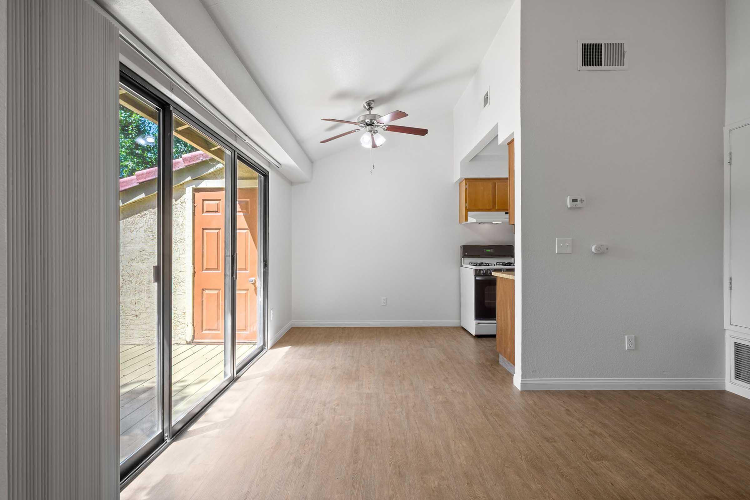 A bright, unfurnished room with large sliding glass doors leading to an outdoor area. A ceiling fan hangs from the ceiling, and there is a kitchen area with wooden cabinets visible in the background. The flooring is light-colored laminate, creating an open and airy atmosphere.