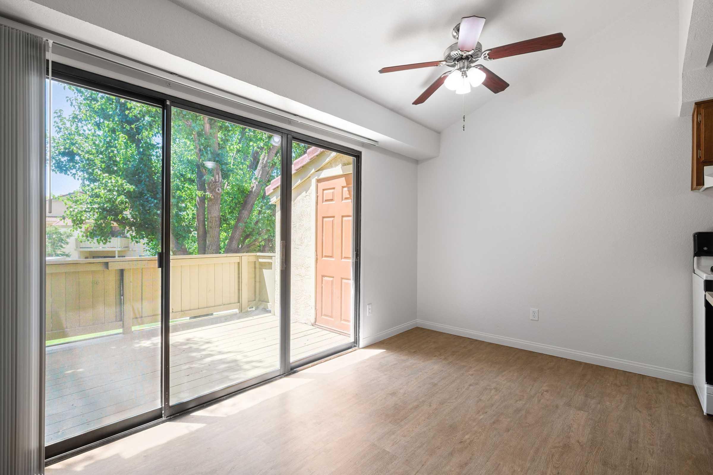 Interior view of a bright room featuring a ceiling fan, large sliding glass doors leading to a deck, and light-colored laminate flooring. There’s a wooden door visible outside, and natural light fills the space, emphasizing its airy atmosphere. The walls are painted in a soft, neutral color.
