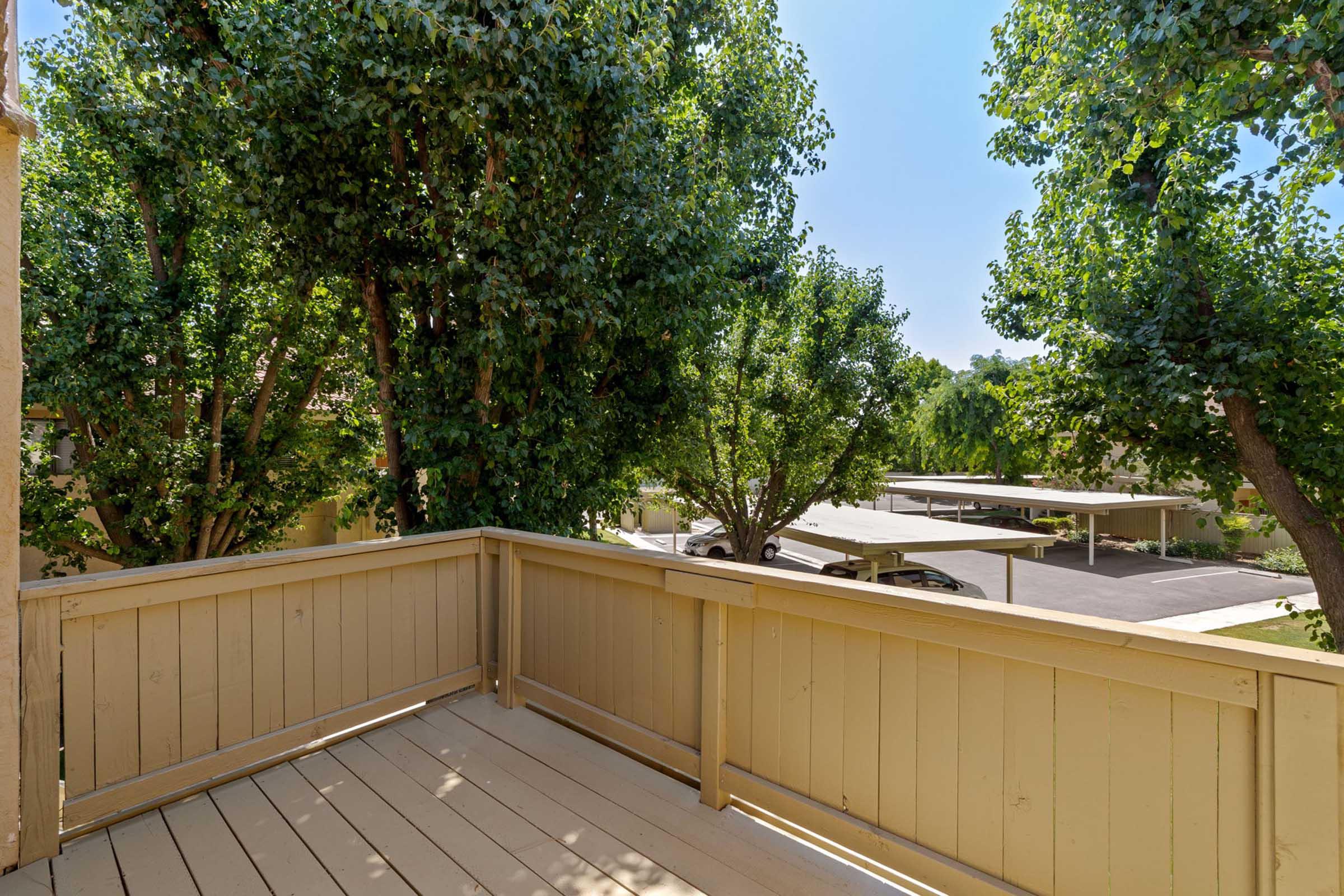 A wooden balcony overlooking a peaceful courtyard with green trees. In the background, there are covered parking spaces and clear blue skies. The scene captures a serene outdoor setting ideal for relaxation.