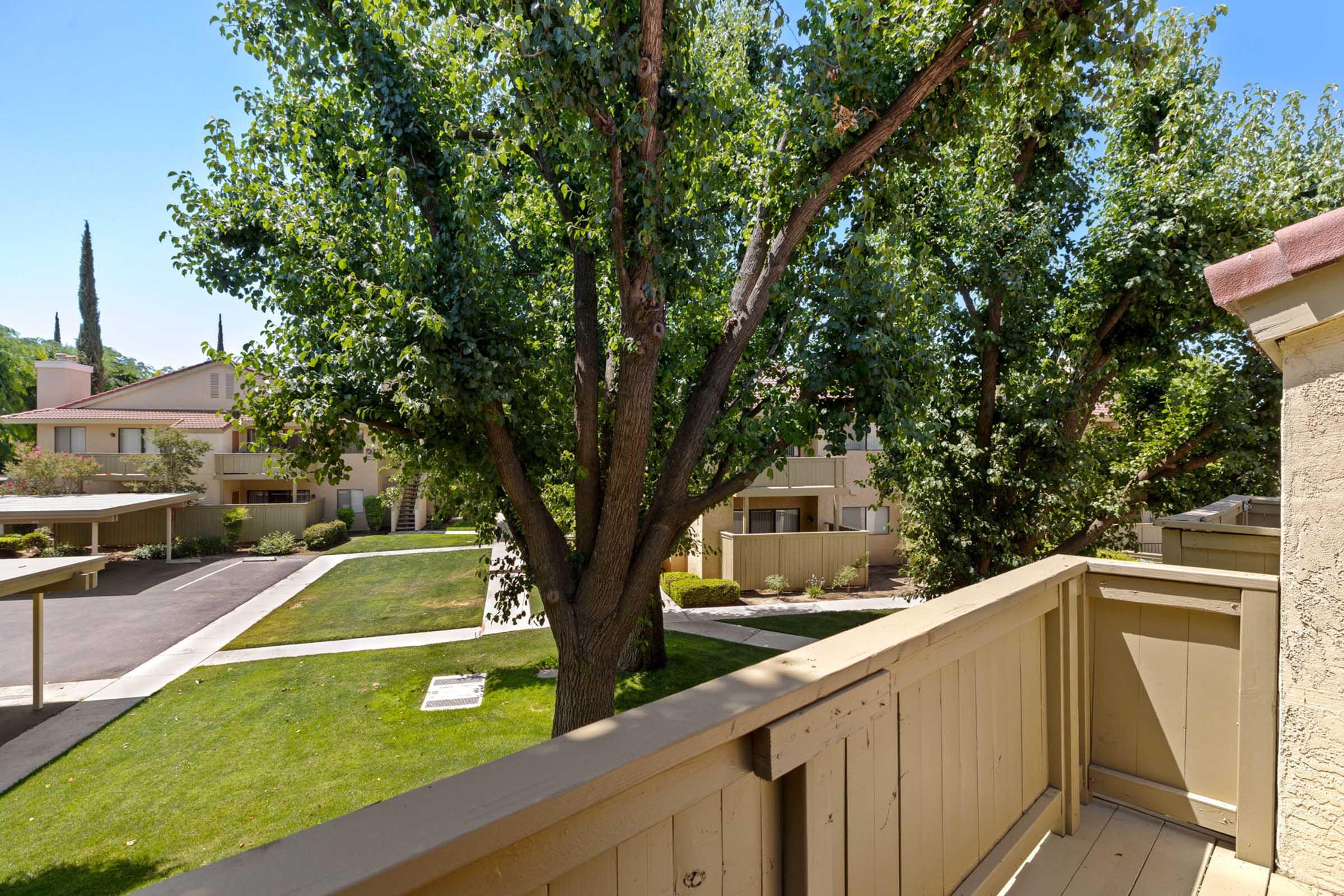 View from a balcony overlooking a landscaped courtyard with green grass and mature trees. In the background, there are residential buildings and parking spaces, all under a clear blue sky. The scene conveys a peaceful and inviting atmosphere.