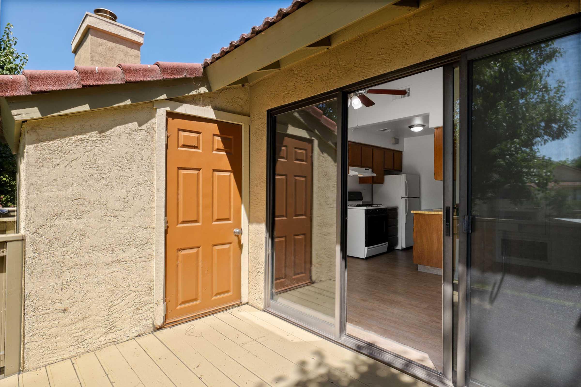 Exterior view of a residential area featuring a patio with a wooden floor, a beige wall, and a brown door. A large glass sliding door provides a view into a well-lit kitchen area with cabinetry and white appliances visible inside. Additional greenery can be seen outside, adding to the serene atmosphere.