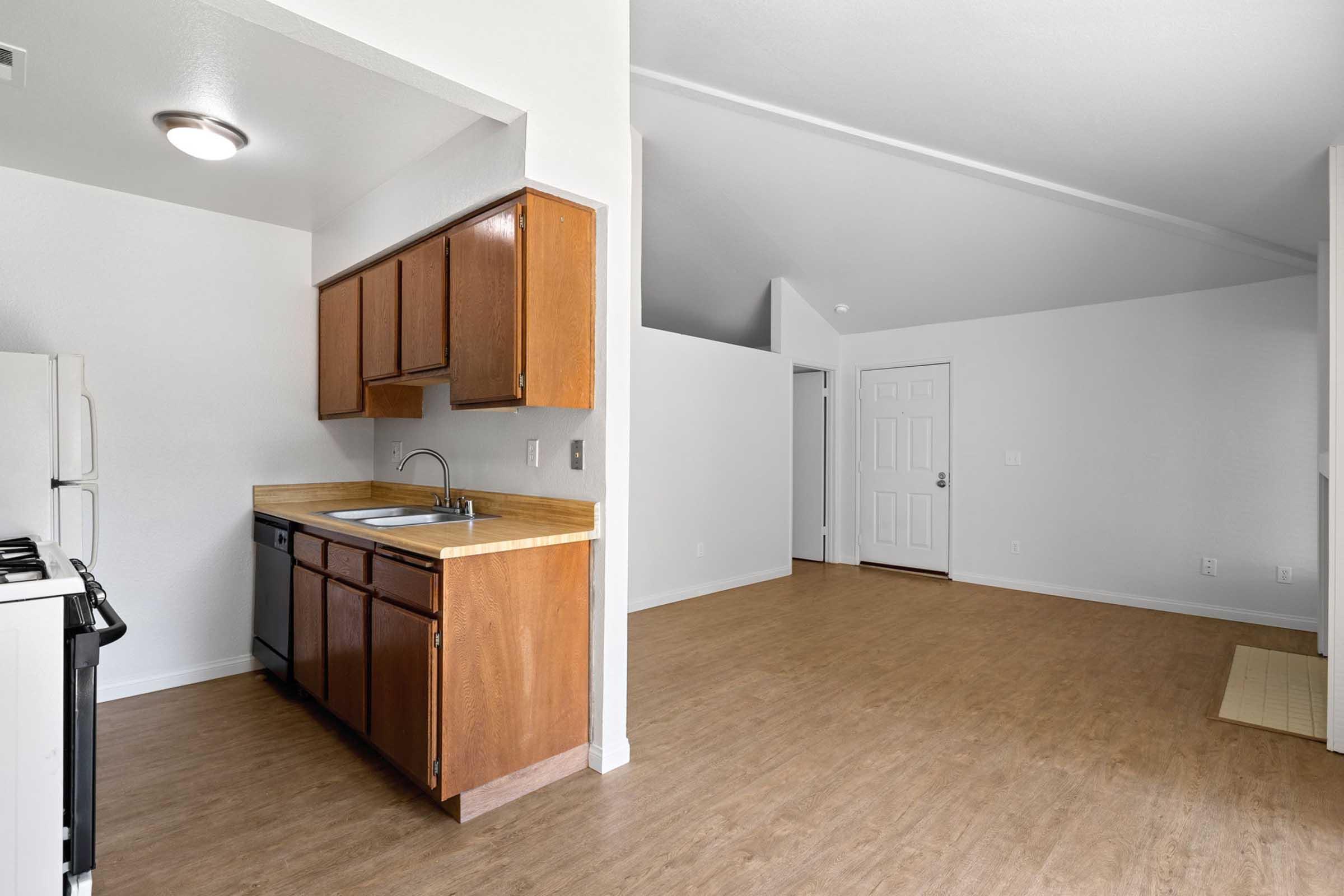 A modern kitchen with wooden cabinets, a sink, and white appliances. The space features a light-colored laminate floor and an open layout leading to a bright, empty living area with white walls and a door. The ceiling is high, creating a spacious feel in the room.