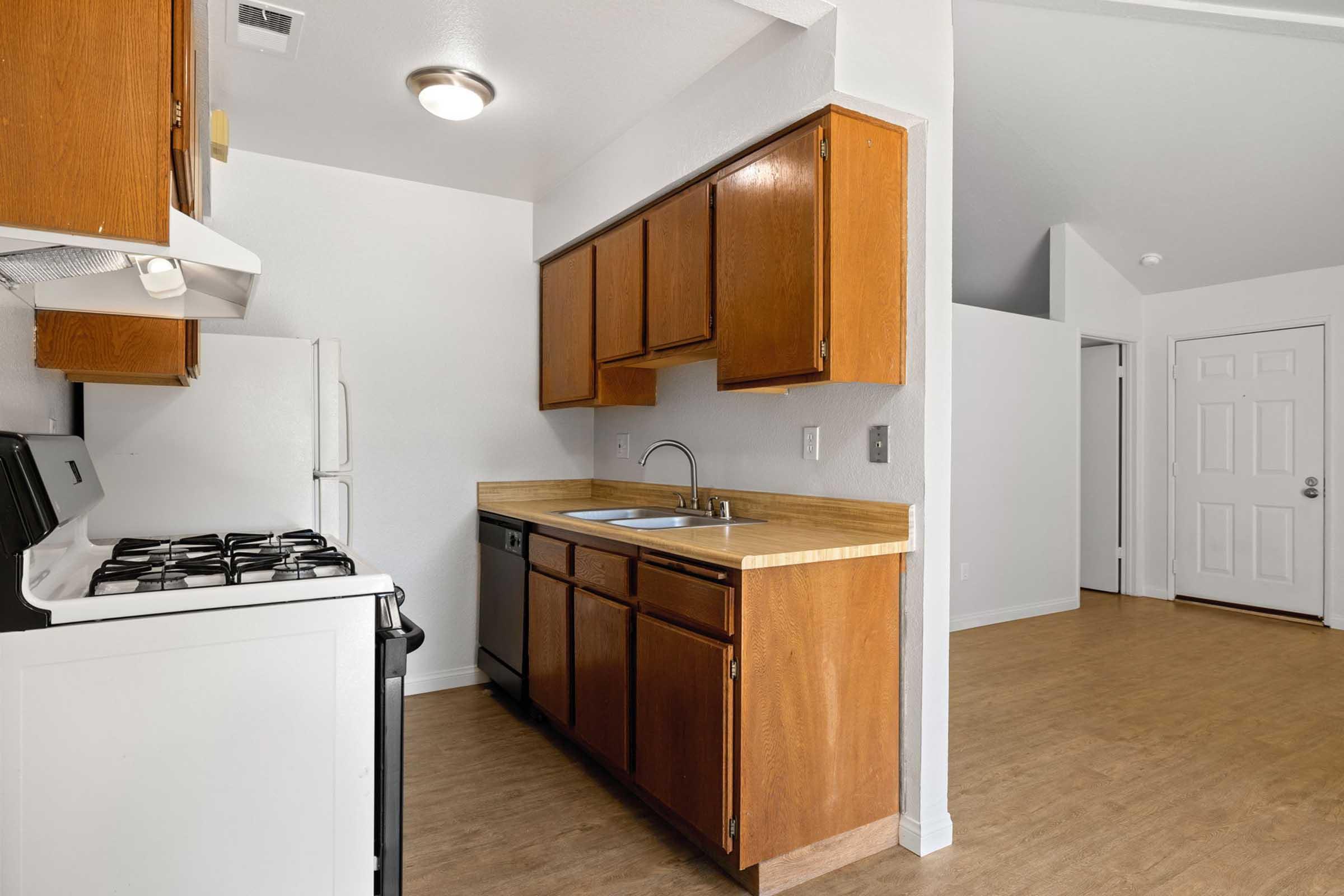 A kitchen with wooden cabinets, a sink, and a white gas stove. There is a dishwasher beside the sink, and the walls are painted white. The floor is light-colored laminate, and there is an open space leading to a living area. A door can be seen in the background.