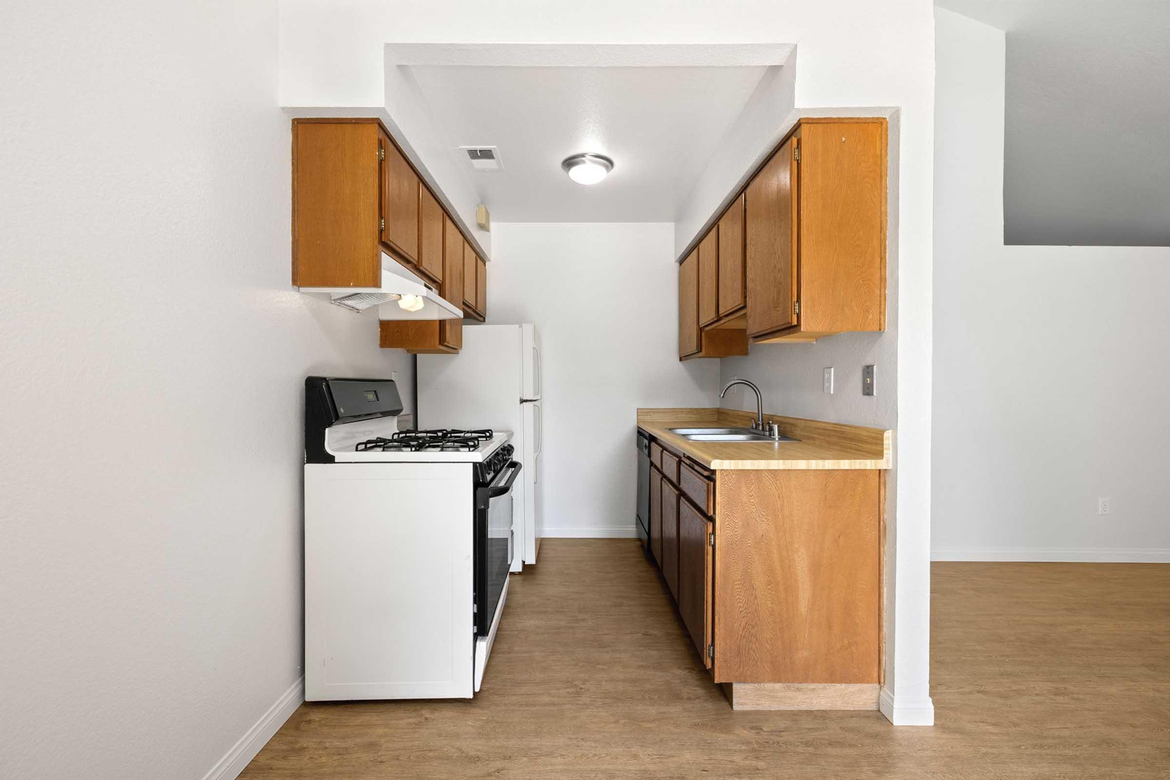 A bright, clean kitchen featuring wooden cabinets, a white gas stove, and a refrigerator. The countertop is beige, and there's a stainless steel sink under a window. The room has light-colored walls and a wood-patterned floor, creating a spacious and inviting atmosphere.