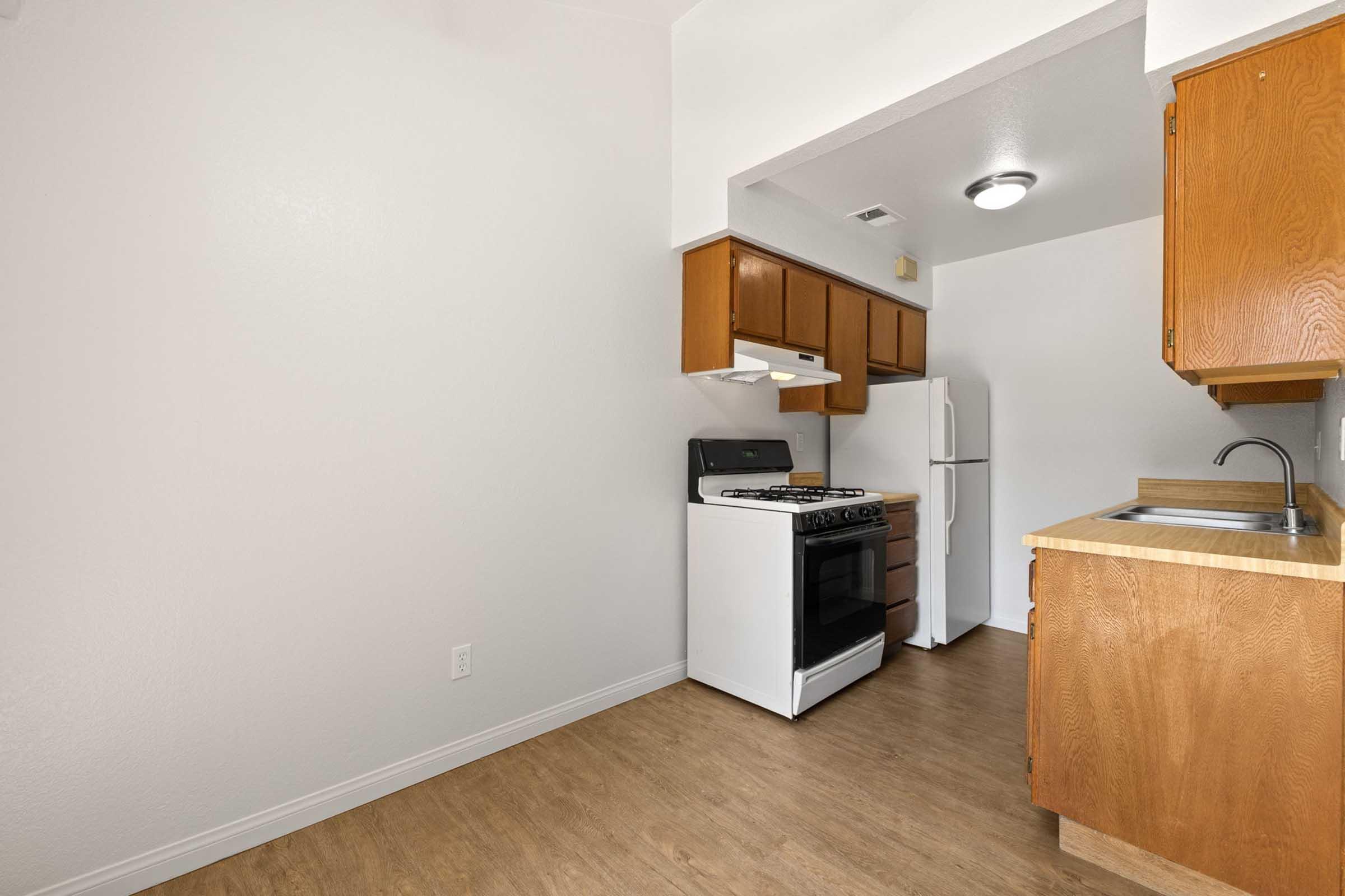 A simple kitchen featuring wooden cabinets, a gas stove, a refrigerator, and a sink. The walls are painted white, and the flooring is light wood. The kitchen is well-lit with a ceiling light, creating a clean and spacious atmosphere.