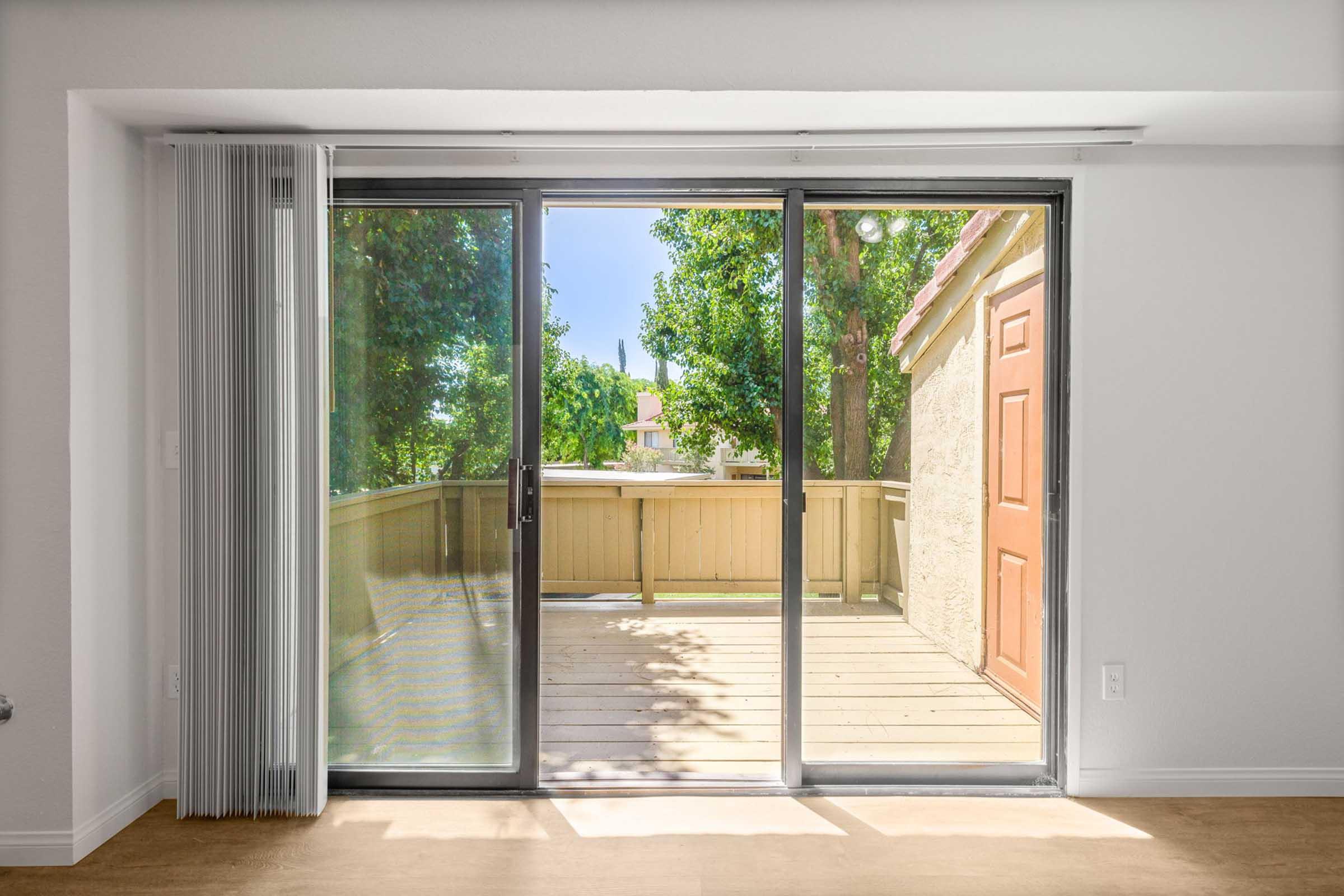Sliding glass doors open to a balcony with wooden flooring, surrounded by greenery. Sunlight streams in through the trees outside, creating a bright and inviting atmosphere. A textured wall and an orange door are visible next to the doors, enhancing the cozy indoor-outdoor connection.