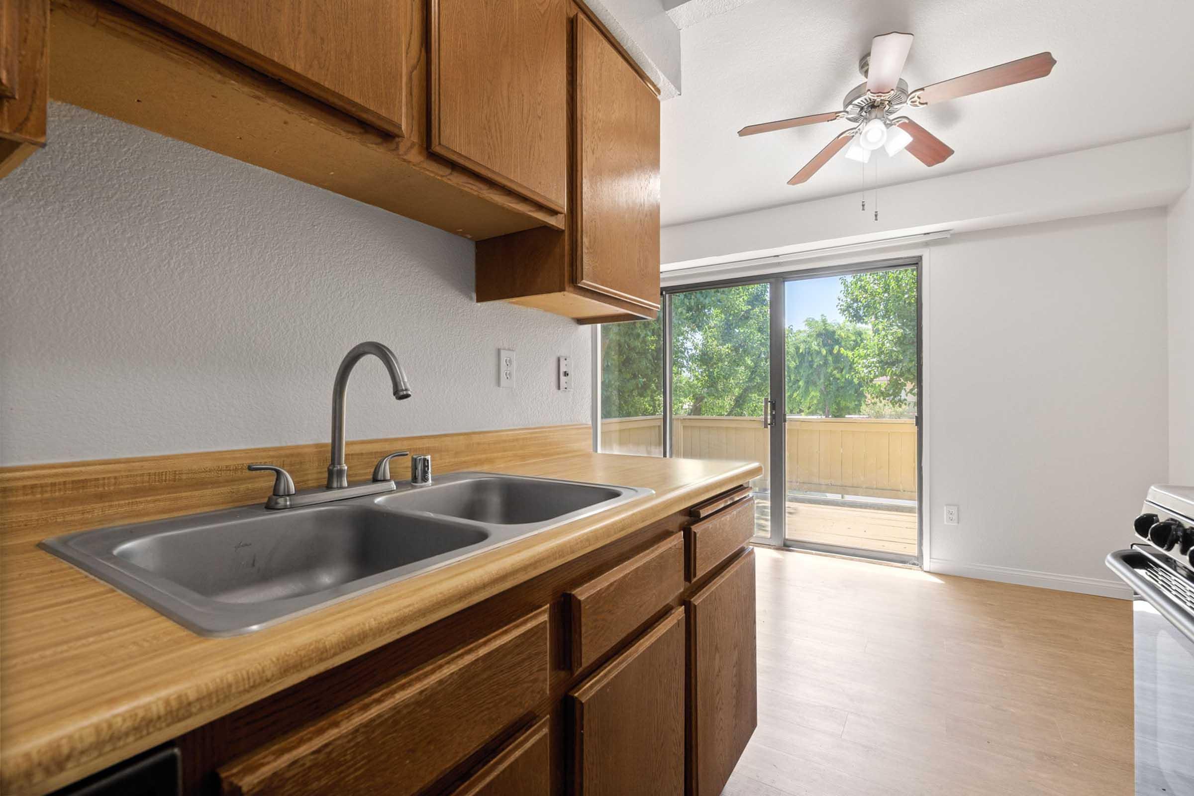 A kitchen with wooden cabinets, a double sink, and a stainless steel faucet. In the background, there is a sliding glass door that leads to a balcony, allowing natural light to enter. A ceiling fan is visible, and the flooring appears to be light-colored, with a glimpse of trees outside.